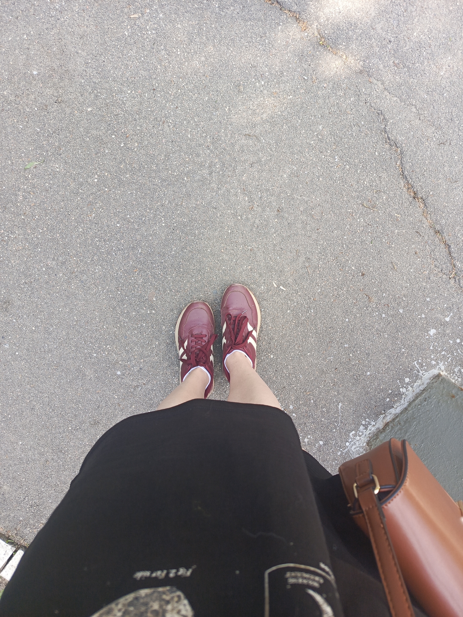 A person is standing on a gray asphalt surface, possibly a sidewalk or street in Dedovsk, Russia, judging by visual cues consistent with urban settings in Russia. The person is wearing maroon sneakers with white stripes, a black skirt, and appears to have a brown leather purse or bag. The asphalt surface has a crack running across it and some minor debris. The person is positioned looking down at their feet.
