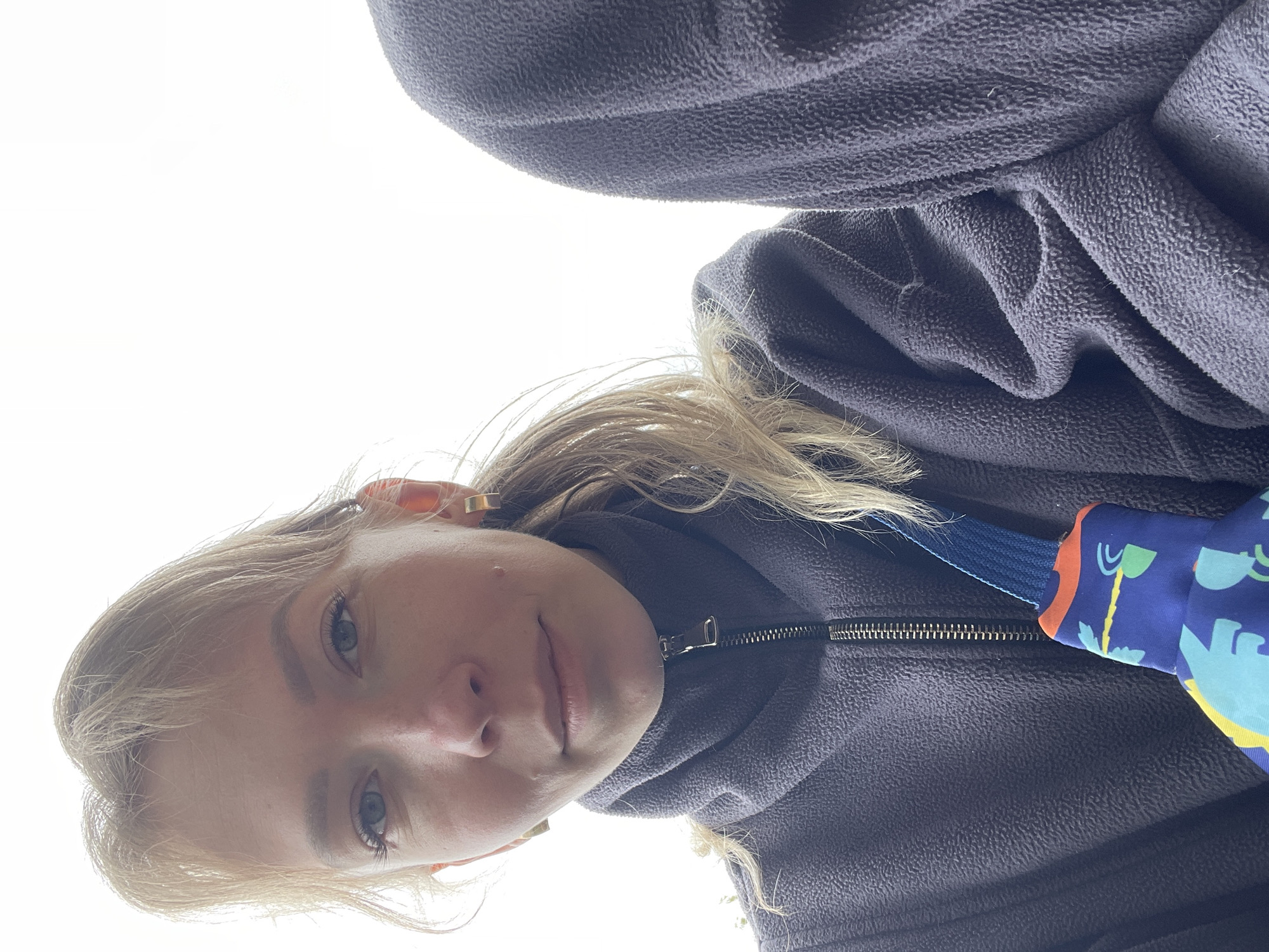 The image presents a close-up, high-angle portrait of a Caucasian woman, taken outdoors, reportedly in the city of Perm, Russia. The woman, with blonde hair styled with bangs and part of it pulled into a ponytail, looks directly at the camera with light-colored eyes and a neutral expression, showing a faint redness on her nose and a small tear-like drop beneath her right eye. She is dressed in a dark grey/purple fleece jacket featuring a high, zipped collar. Gold-colored hoop earrings are visible on both ears. A small, vibrant blue pouch adorned with multi-colored cartoon-like patterns, attached to a blue strap, rests on her left chest area. To the right, a segment of another person or object covered in similar dark grey/purple fleece is partially visible. The background is an undifferentiated, overexposed white sky, illuminated by bright overhead natural light.