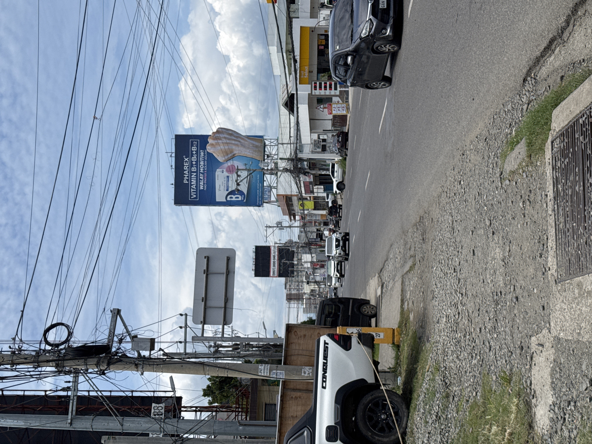 This media file captures a daytime urban street scene in Davao City, Philippines, viewed from a slightly elevated perspective beside the road.
In the foreground, a white pickup truck, emblazoned with "CONQUEST" on its side, is parked or stopped on a gravelly area next to the asphalt road. A yellow post marked "27" stands nearby. Immediately to the left, a dense tangle of overhead utility wires and cables crisscrosses the sky, anchored to a concrete pole bearing signs like "ELCO" and partial phone numbers (e.g., "099984", "0932670").
The road features white lane markings, with a black car visible in motion or parked. Beyond it, a building with a yellow "Select" sign is discernible. Further down the street, additional vehicles suggest ongoing traffic. A large billboard prominently displays "PHAREX® VITAMIN B1+B6+B12" with the phrases "WALAY MOBITIW!" (meaning "Will not give up!") and "MAG-PHAREX B ADLAW-ADLAW!" (meaning "Take Pharex B every day!"). The billboard also features a graphic of a human fist.
In the background, a street lined with various commercial buildings extends into the distance. Another large black billboard is visible, featuring a phone number: "(0917) 3000-840." The sky above is partly cloudy, indicating clear daylight conditions. The overall scene depicts a typical bustling street environment in a Philippine city, characterized by commercial activity and extensive overhead wiring.
