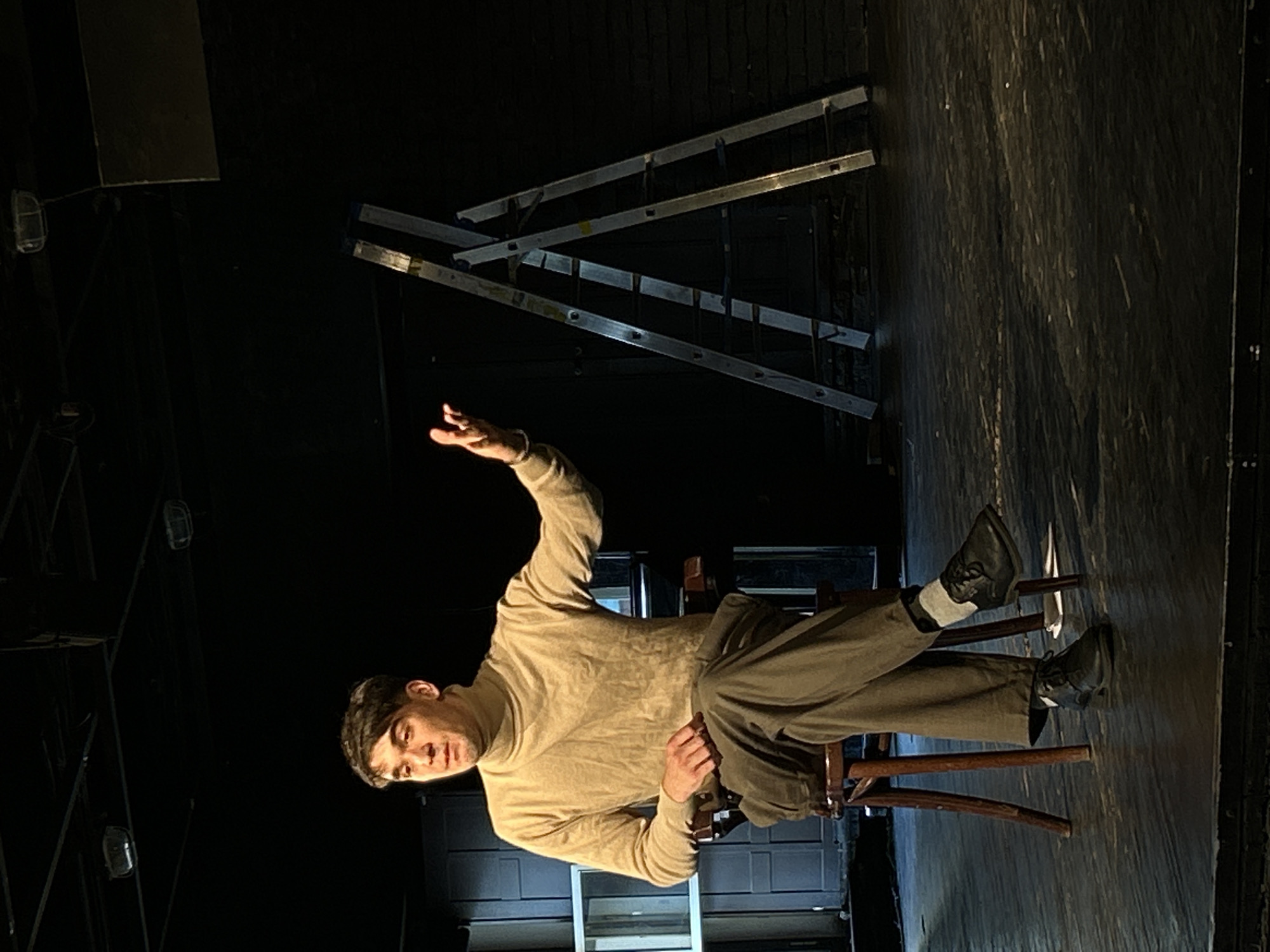 A man is seated on a wooden chair in what appears to be a dimly lit stage or rehearsal space. He is positioned slightly off-center to the left, facing towards the right side of the frame. He wears a light-colored (beige or tan) turtleneck sweater, dark grey trousers, white socks, and black dress shoes. His right arm is raised and bent at the elbow, with his hand open and palm facing upwards, in a gesture that could signify questioning, offering, or reaching. His left hand rests on his left thigh. He looks slightly upwards and to his left (from his perspective) with a focused or contemplative expression.
Behind the man, several sections of metal ladders are leaning against a dark backdrop, creating a diagonal pattern in the upper right portion of the image. The floor beneath him is dark wood, likely part of a stage. The lighting is low-key, with a subtle highlight on his raised hand and face, suggesting intentional stage lighting. The overall atmosphere is quiet and somewhat dramatic, possibly indicating a theatrical performance or rehearsal. No visible text is present in the image.