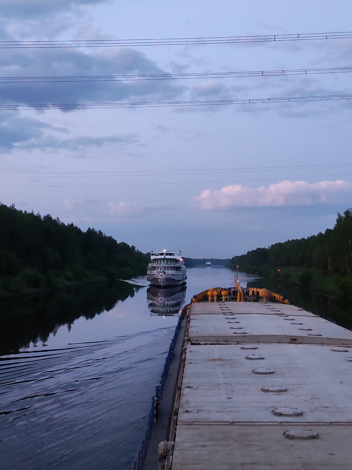 The image shows a waterway in Zaprudnya, Russia. A large barge occupies the foreground, with a white deck featuring numerous circular hatches. A passenger riverboat is visible in the middle distance, moving towards the viewer, with its reflection visible in the water. Dense green trees line the banks of the waterway on both sides. Overhead, electrical power lines stretch across the frame against a twilight sky. The lighting suggests it is dusk or dawn. The waterway is calm, with only minor disturbances visible from the boats' movement. The barge and riverboat are navigating the waterway.