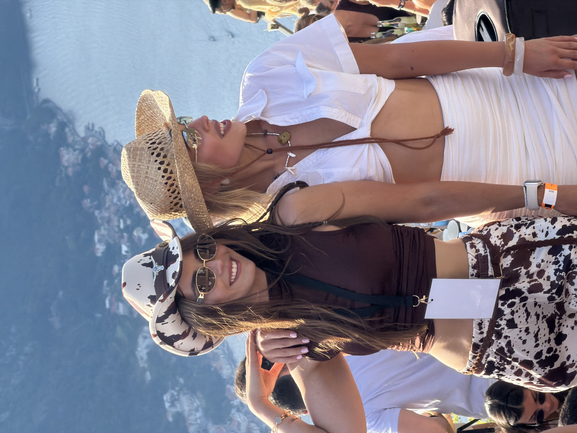 Two young women in cowboy hats and sunglasses are standing on an elevated viewpoint overlooking the city of Kotor, Montenegro. The woman on the left wears a brown sleeveless top and cow-print pants, with a white lanyard and tag. The woman on the right wears a white tied-front shirt and a white ruched skirt. Both are smiling and appear to be interacting. The background shows a body of water and a hilly landscape with buildings. The lighting suggests it is daytime, likely late afternoon or early evening due to the warm tones.