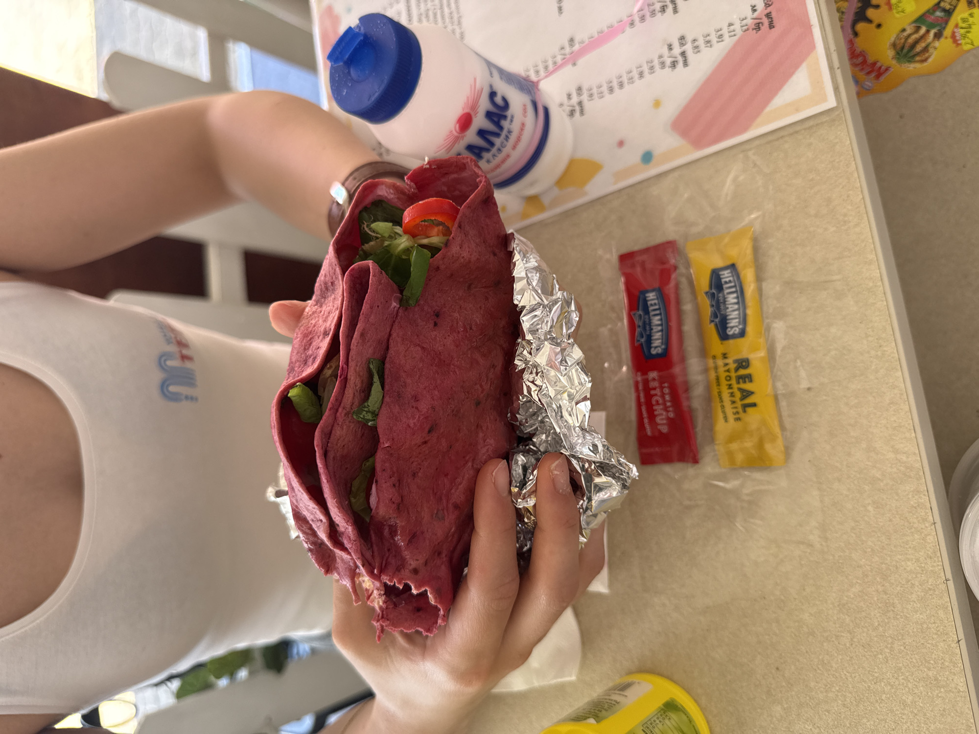 In this close-up, top-down shot, a person is holding a vibrant pink wrap. The wrap, which appears to be made from a beetroot tortilla, is filled with fresh green lettuce, sliced red tomatoes, and possibly other vegetables. It is partially wrapped in crinkled aluminum foil. Next to the wrap, two small packets of Hellmann's condiments are placed: one red packet labeled "TOMATO KETCHUP" and a yellow packet labeled "REAL MAYONNAISE". In the background, a blue-capped bottle of "ALAS KLASIK" drink is visible, along with a menu showing prices in Bulgarian Leva. The setting appears to be outdoors on a table, suggested by the natural lighting and the presence of what looks like an outdoor seating area in the blurred background. The overall impression is of a casual meal, perhaps lunch, on a sunny day. The focus is on the colorful and appetizing wrap, highlighting its fresh ingredients and the accompanying sauces.