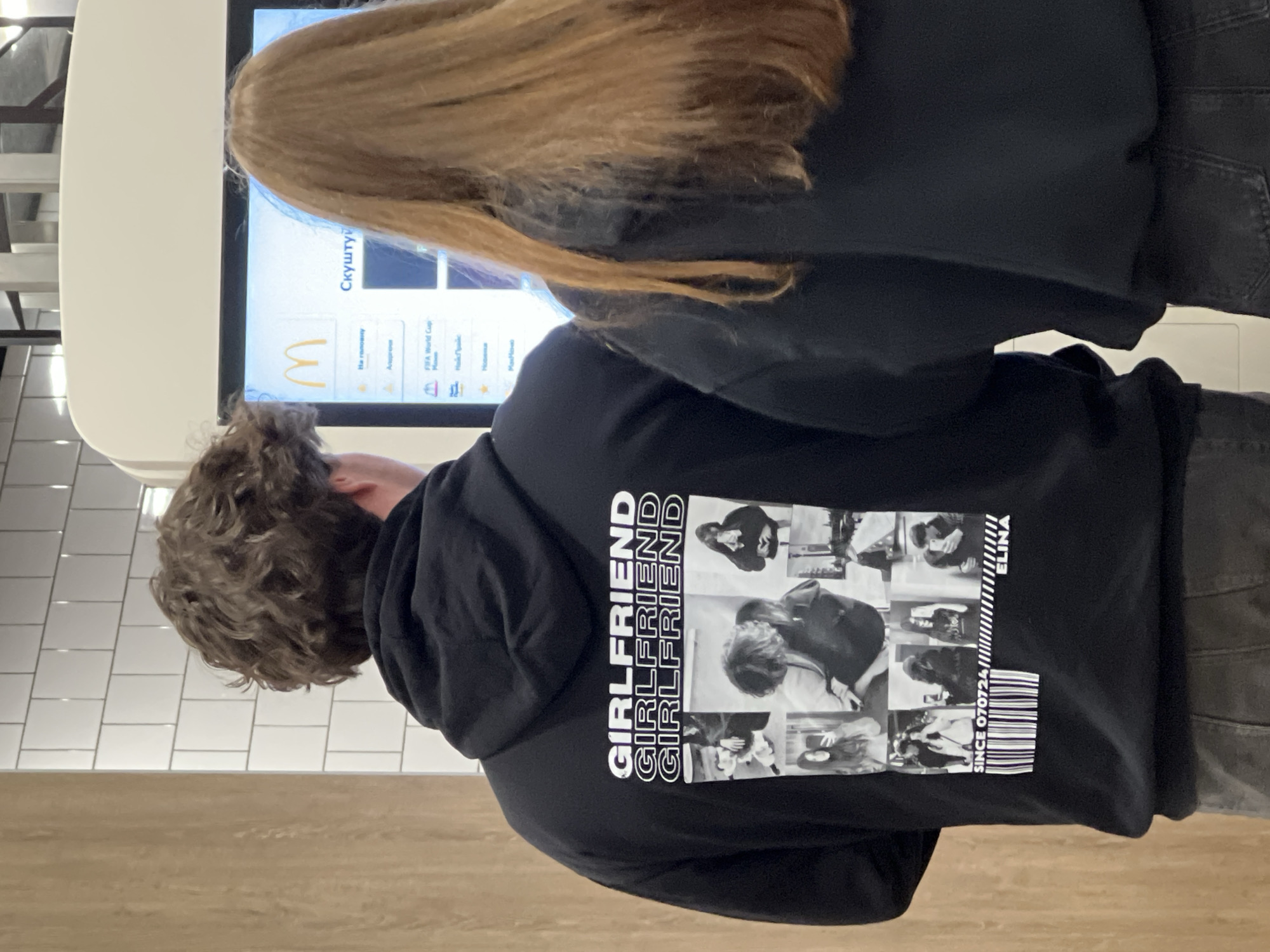 Two individuals, a male with curly brown hair and a female with long auburn hair, stand in front of a McDonald's self-order kiosk. The male is wearing a black hoodie with a graphic print on the back that includes the text "GIRLFRIEND" repeated three times, followed by a collage of black and white photographs and a barcode. The female is wearing a black hoodie and is facing the kiosk screen, which displays the McDonald's logo and various menu items in what appears to be Russian or Ukrainian. The setting is indoors, likely within a McDonald's restaurant in Ternopil, Ukraine, indicated by the kiosk's interface and common McDonald's interior design elements like white tiled walls and a wooden accent wall. The interaction between the two appears to be one of ordering food, with the female presumably navigating the kiosk.
