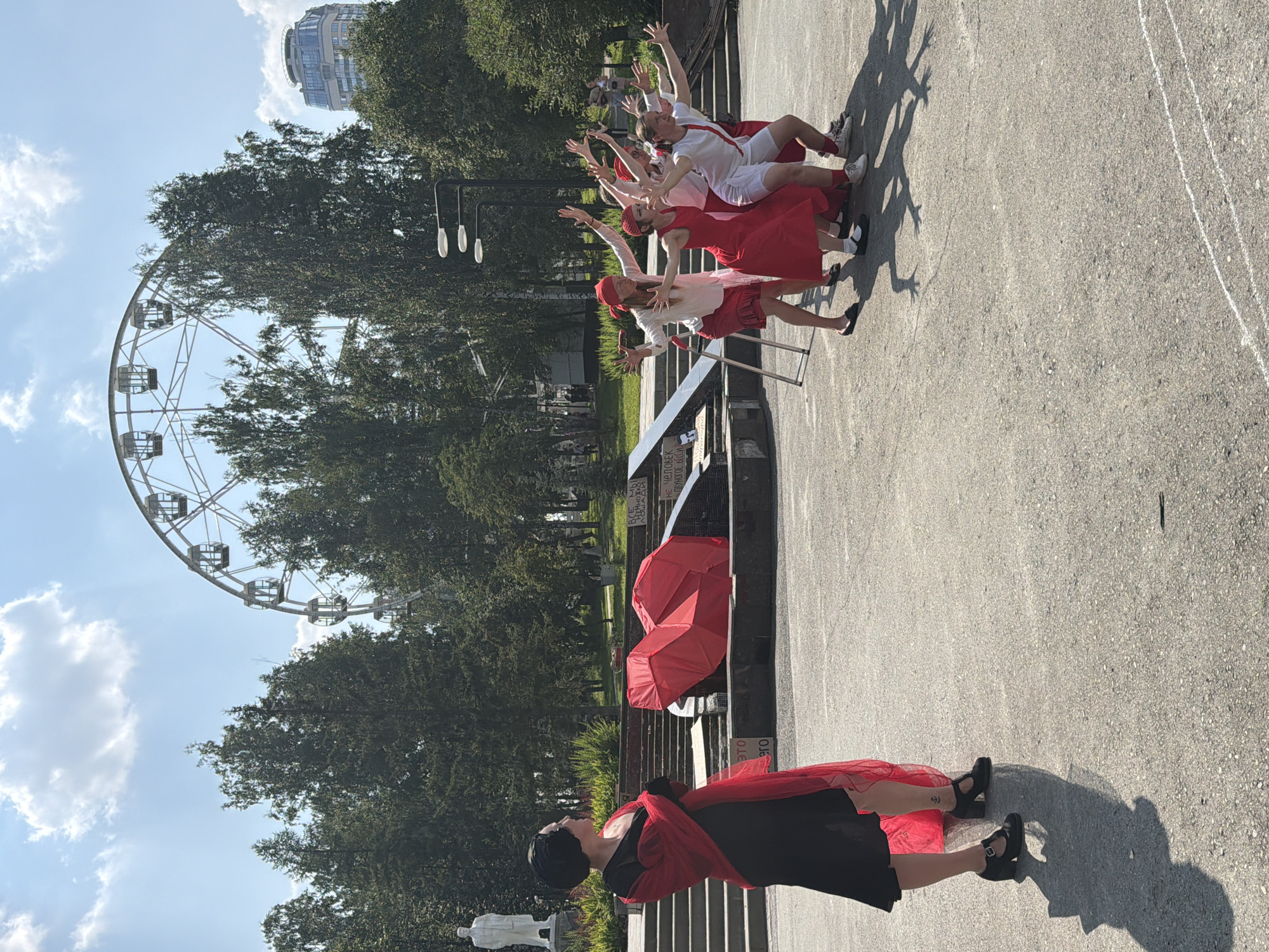 In Yekaterinburg, Russia, a group of performers in red and white attire are captured in a synchronized pose outdoors, with their arms extended. They are positioned on a paved surface in front of a large red, heart-shaped sculpture. To the left, a woman in a black dress and a red shawl stands facing away from the camera, observing the group. Behind the performers and the sculpture, a white Ferris wheel is visible in the background, suggesting a park or amusement area. Lush green trees line the perimeter of the scene, and a tall, modern building with a helipad can be seen in the distance, indicating an urban setting. The sky is blue with scattered white clouds, and the sunlight casts long shadows on the ground.