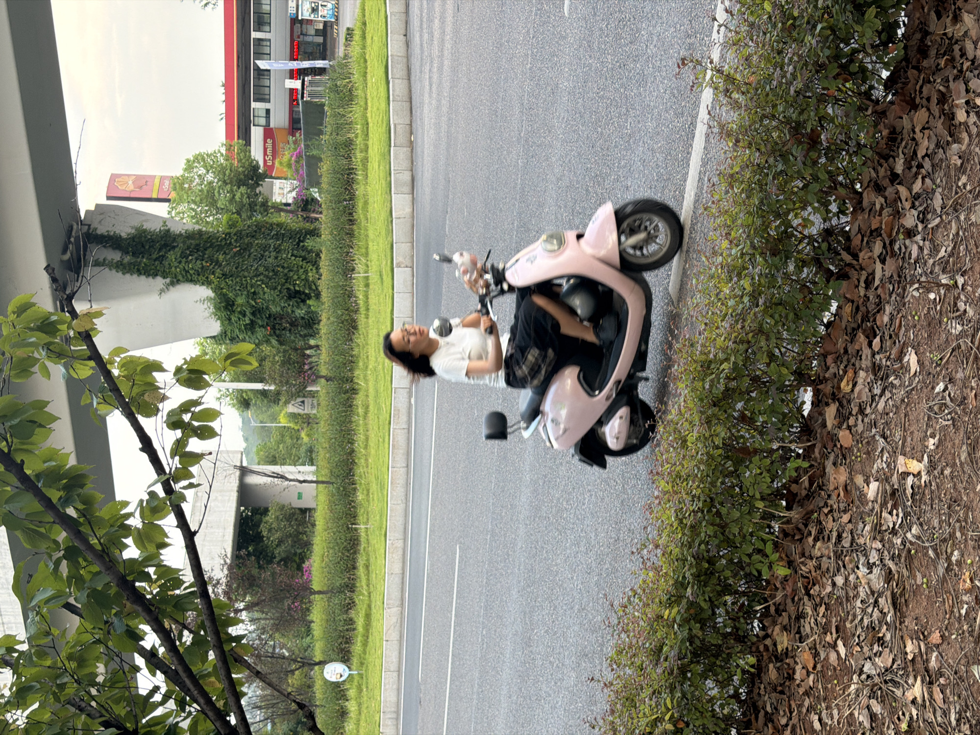 A woman is riding a pink electric scooter on a paved road in Jiangjin, China. She is wearing a white t-shirt and a plaid skirt, and has dark hair and glasses. The scooter is moving from left to right. In the background, there is a highway overpass, a gas station with a Shell logo, and lush green vegetation. The foreground consists of a row of manicured bushes and fallen leaves on the ground. The sky is overcast.