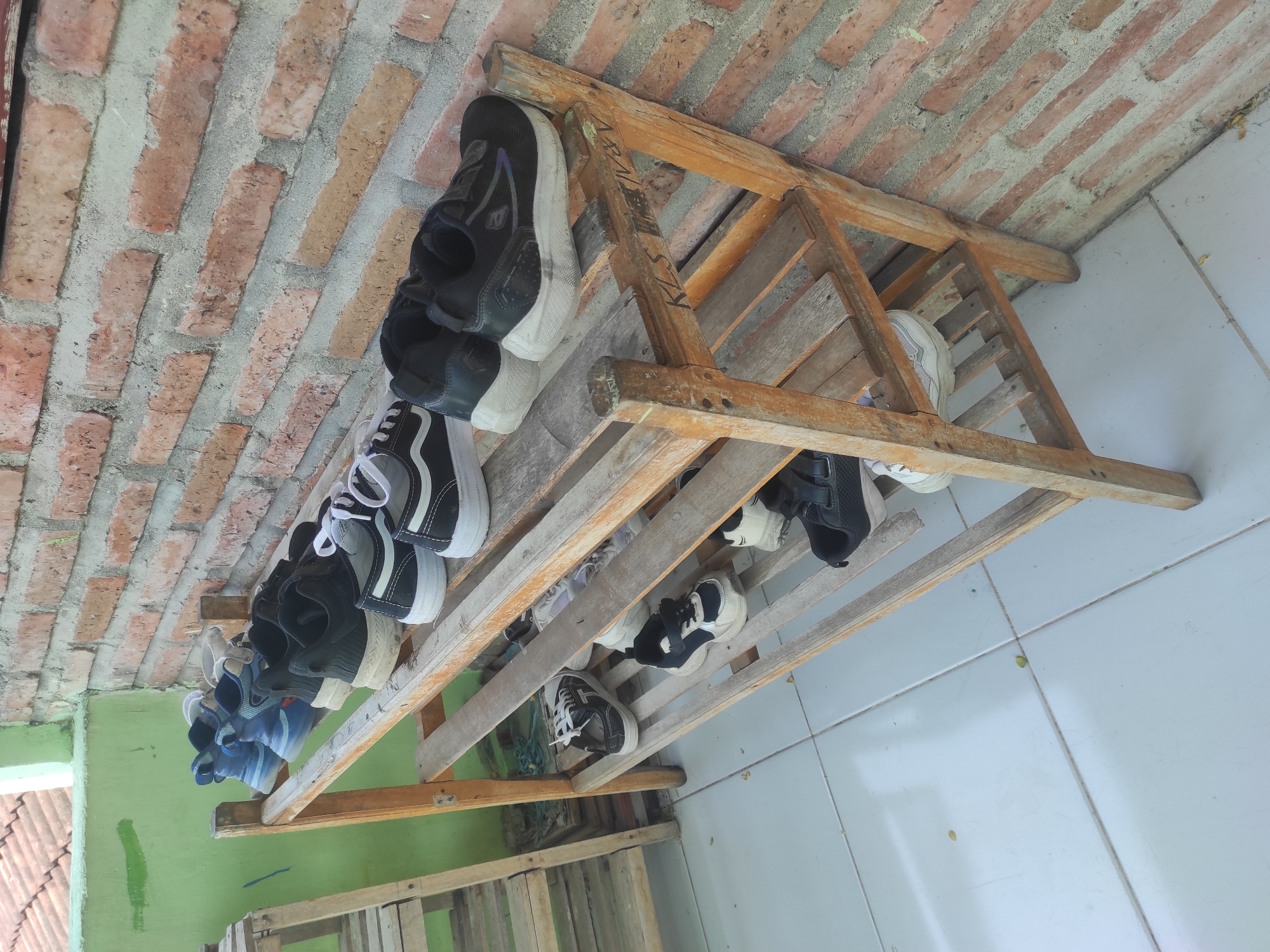 This image depicts a three-tiered wooden shoe rack situated outdoors, leaning against a brick wall. The shoe rack is filled with various pairs of shoes, primarily sneakers. On the top shelf, there are several pairs of blue and black sneakers. The middle shelf holds more black and white sneakers, including a pair with white laces. The bottom shelf contains black and white sneakers, with one pair being black with blue accents and another pair being mostly white. The environment appears to be a residential or communal outdoor space, with a light-colored tiled floor and a green painted wall visible to the left. The brick wall shows signs of age and weathering. The overall scene is a mundane, everyday arrangement of footwear. The location is identified as the city of Klepek, Indonesia, suggesting a setting in Southeast Asia.