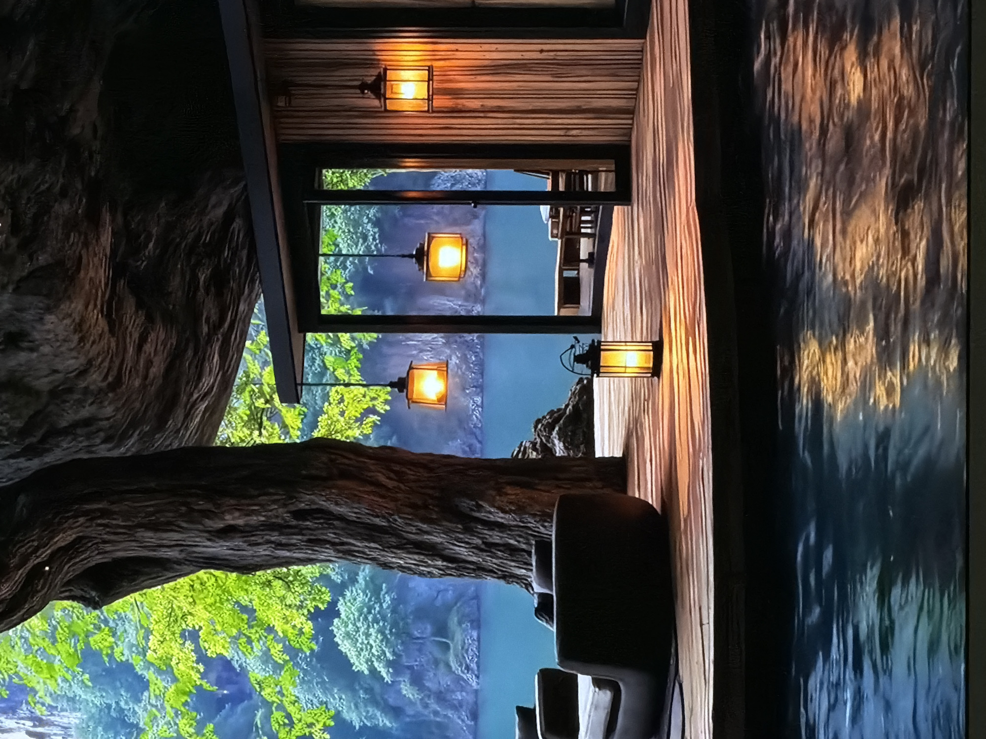 This media file captures an immersive interior setting, possibly a themed restaurant or lounge, designed to resemble a treehouse or forest retreat. The image is oriented vertically but depicts a horizontally structured scene.
In the foreground, a warm, inviting interior is visible, characterized by wooden paneling on the walls. Several antique-style lanterns, both wall-mounted and hanging, cast a soft, orange glow, illuminating the space. The light sources include a wall-mounted lantern at the top, a hanging lantern in the center, and another wall-mounted lantern on the right side. On the right, a textured surface reflects light with ripple-like patterns, suggestive of flowing water or a simulated waterfall. Dark, comfortable-looking chairs or seating are partially visible in the lower right, implying a dining or relaxation area.
Dominating the left side of the interior are large, textured tree trunks, giving the impression of being nestled within a forest. Through a large, rectangular opening or screen, a vibrant "outdoor" scene is displayed. This simulated landscape features lush green foliage and trees, with a bright blue body of water or sky extending into the distance. The contrast between the warm, dim interior lighting and the brightly lit exterior view suggests the "outdoor" scene might be a digital projection or a large window overlooking an elaborately designed diorama.
No people are visible, and no specific activity is taking place; the scene is static. The warm artificial lighting inside creates a cozy ambiance, while the depicted "daytime" exterior enhances the naturalistic theme. There is no visible text in the image. The location context provided is Sofiivska Borshchahivka, Ukraine.