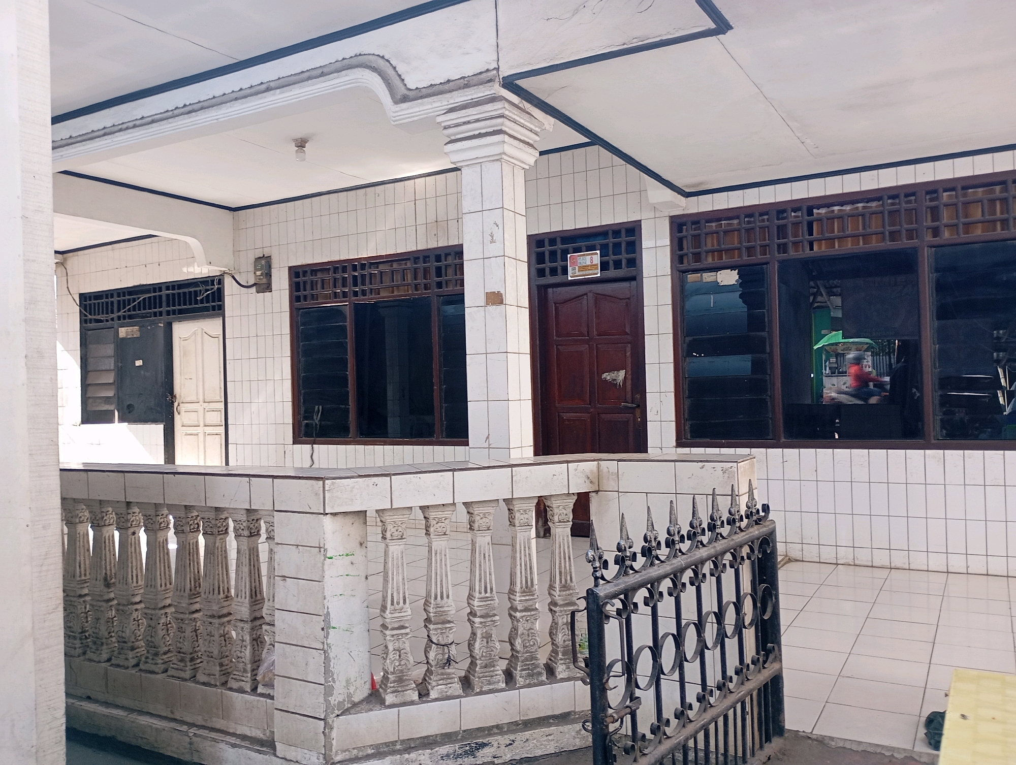 The image depicts the exterior of a building, possibly a house or a small shop, in Babelan, Indonesia. The building features white tiled walls, dark-framed windows with horizontal louvers, and a covered patio area. A white concrete balustrade with decorative pillars runs along the edge of the patio, and a black metal fence is visible in the foreground. There is a wooden door with a sign on it, and reflections in the windows suggest activity inside or a view of the street. The lighting indicates it is daytime, and the overall impression is one of a modest, functional building in a residential or commercial area.