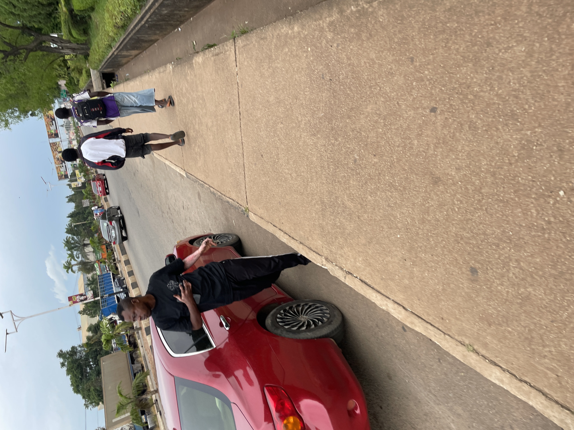 The image depicts a street scene in Jos, Nigeria. In the foreground, a red car is parked on the left side of the road. A young man in a black t-shirt and black track pants stands next to the car, holding a phone in his right hand. His left arm is bent at the elbow, with his hand near his chest. Further down the sidewalk, two young men are walking away from the camera. The man on the left wears a white and red jacket and dark shorts, while the man on the right has a backpack, a purple and white shirt, and light blue wide-leg pants. The environment includes a paved sidewalk, a road with other vehicles in the distance, trees lining the right side of the street, and buildings and billboards in the background. The sky is overcast.