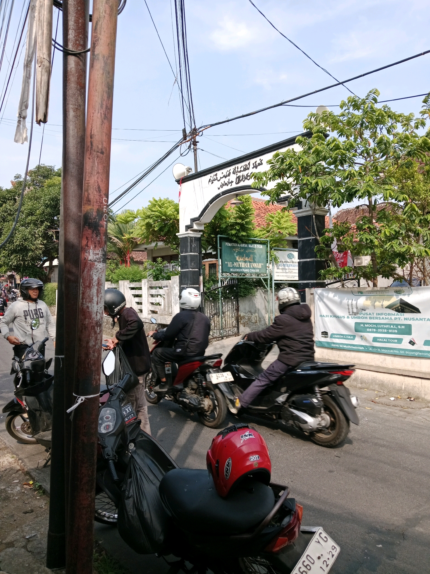 The image shows a street scene in Bojongherang, Indonesia. Several people are on or near motorcycles. In the foreground, a person wearing a grey hoodie and a helmet stands next to a black motorcycle. To their left, another person wearing a black helmet and jacket is leaning over a black motorcycle. Further back, a person in a white helmet and jacket rides a red motorcycle. The background shows a building with an archway and Arabic inscriptions, likely a mosque or religious institution, with trees and power lines overhead. A banner with text in Indonesian, including "PUSAT INFORMASI HAJI KHUSUS & UMROH..." and a phone number, is visible on the right side. The overall environment suggests a sunny day on a street with modest buildings and vegetation.