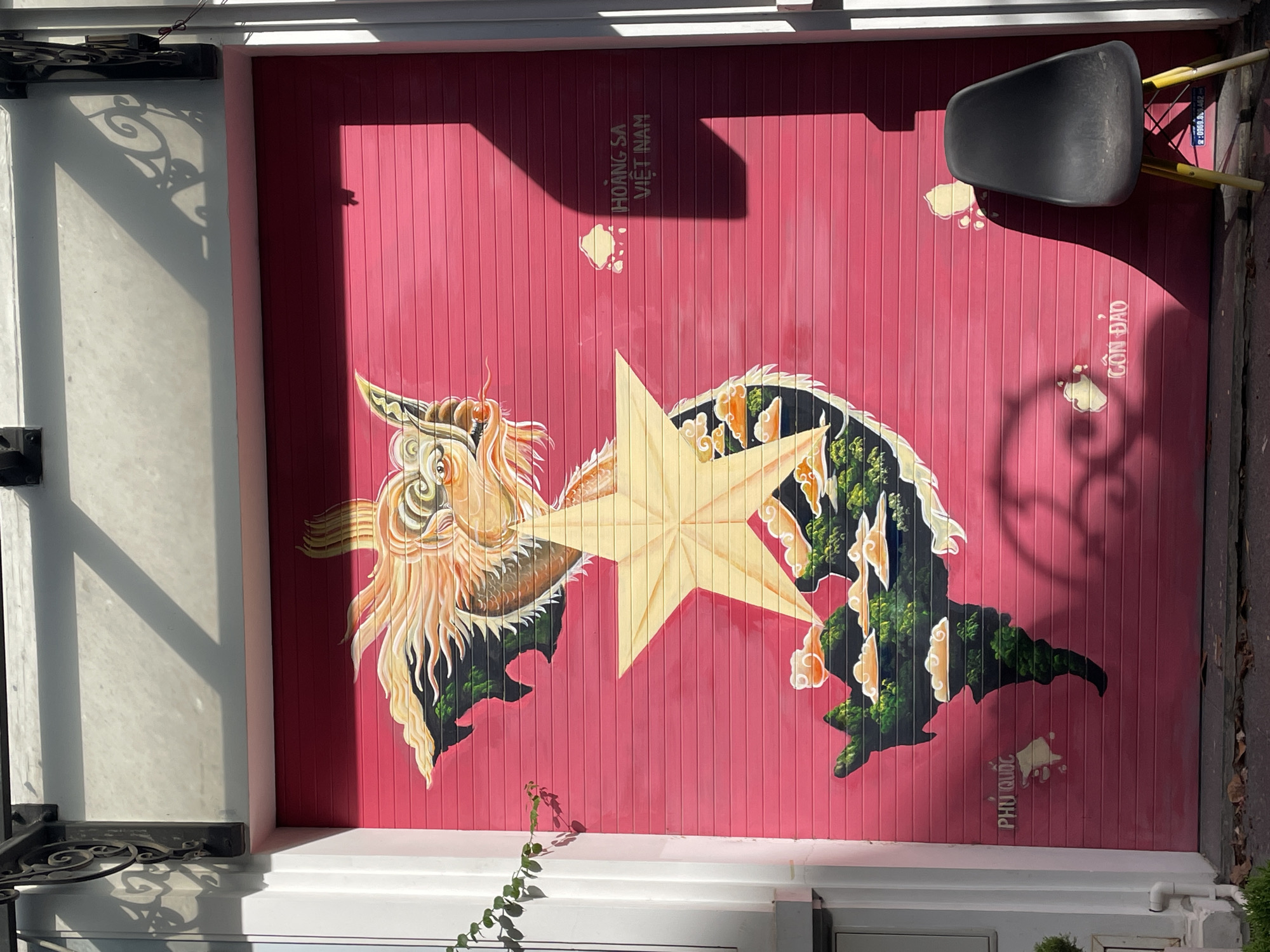 This image displays a vibrant mural painted on a bright pink roller shutter door. The mural depicts a stylized map of Vietnam, superimposed with a yellow star, and a dragon's head at the top. Islands are represented as green land masses with clouds. Text labels for "Phú Quốc" and "Côn Đảo" are visible on the map. To the right, text reads "HOÀNG SA VIỆT NAM". A dark gray chair with yellow legs stands in front of the shutter. Sunlight casts strong shadows across the scene, indicating it is outdoors. The location is identified as Hanoi, Vietnam.