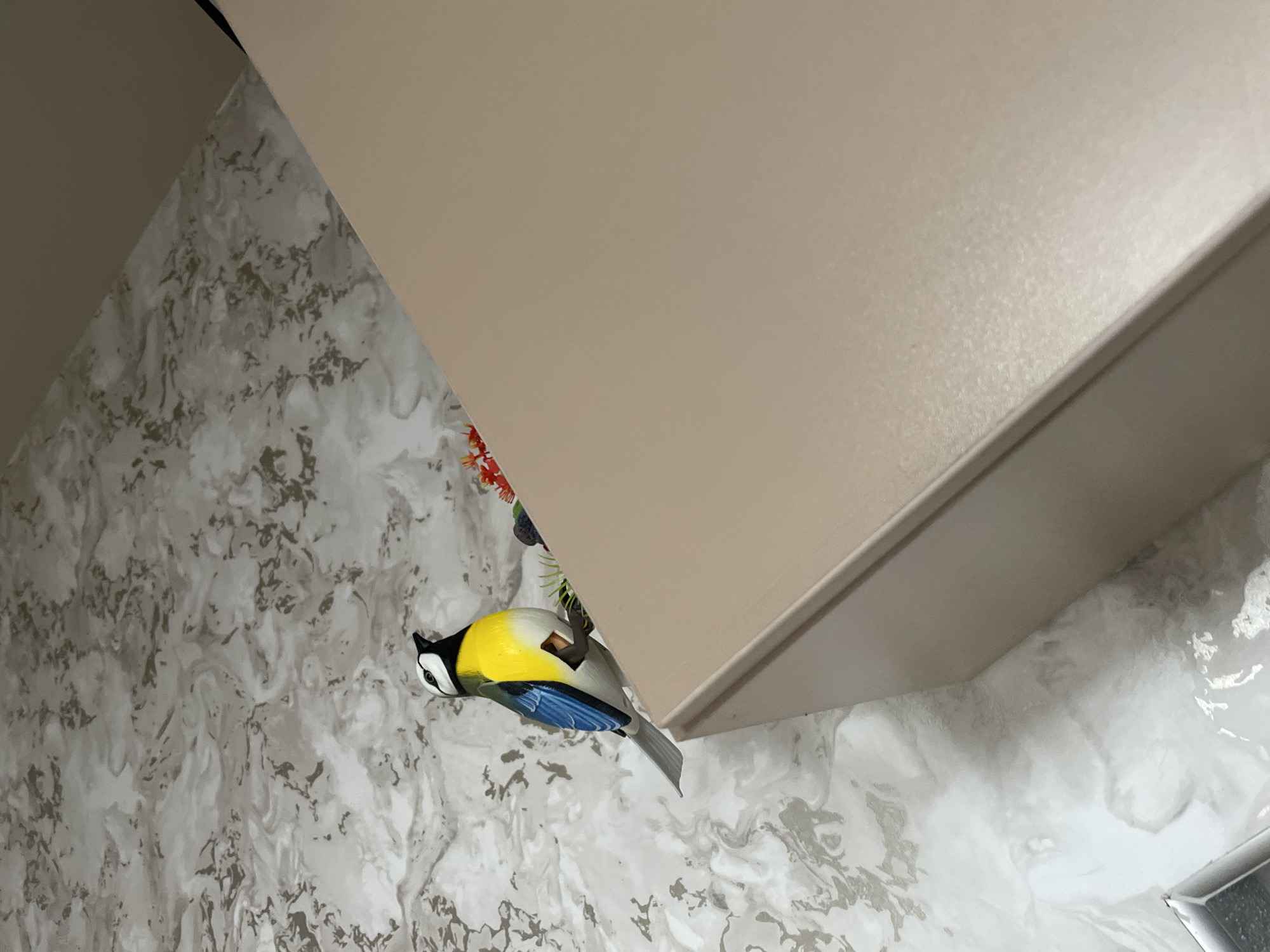 The image shows a Eurasian blue tit perched upside down on a shelf or cabinet. The shelf is a light tan color. The wall behind the bird is covered in a gray and white marble-patterned wallpaper. A small green and red decorative piece is also visible near the bird. The location is not discernable from the visual information provided.
