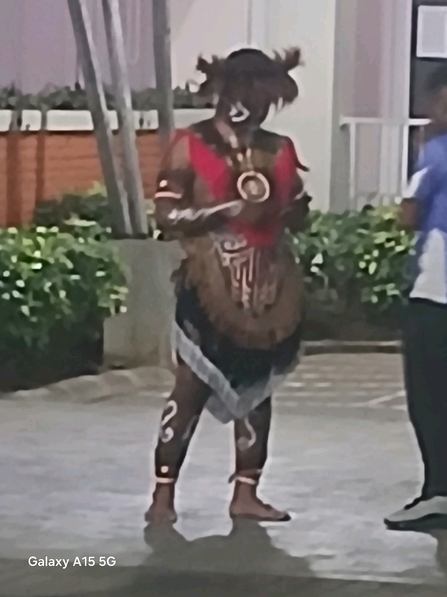 The image shows a person in traditional attire, possibly a dancer or performer, standing outdoors. The person is wearing a headdress with feathers, face paint, a red and brown garment, and intricate patterns on their legs. They are holding an object in their hands. To the right, another person in a blue and white shirt is partially visible. The background includes lush green bushes, a brick wall, and some vertical structures. The ground appears to be paved. The overall setting suggests an outdoor event or performance. The text "Galaxy A15 5G" is visible in the lower-left corner, indicating the device used to capture the image. There is no specific information within the image that allows for identification of the location as Surabaya, Indonesia.
