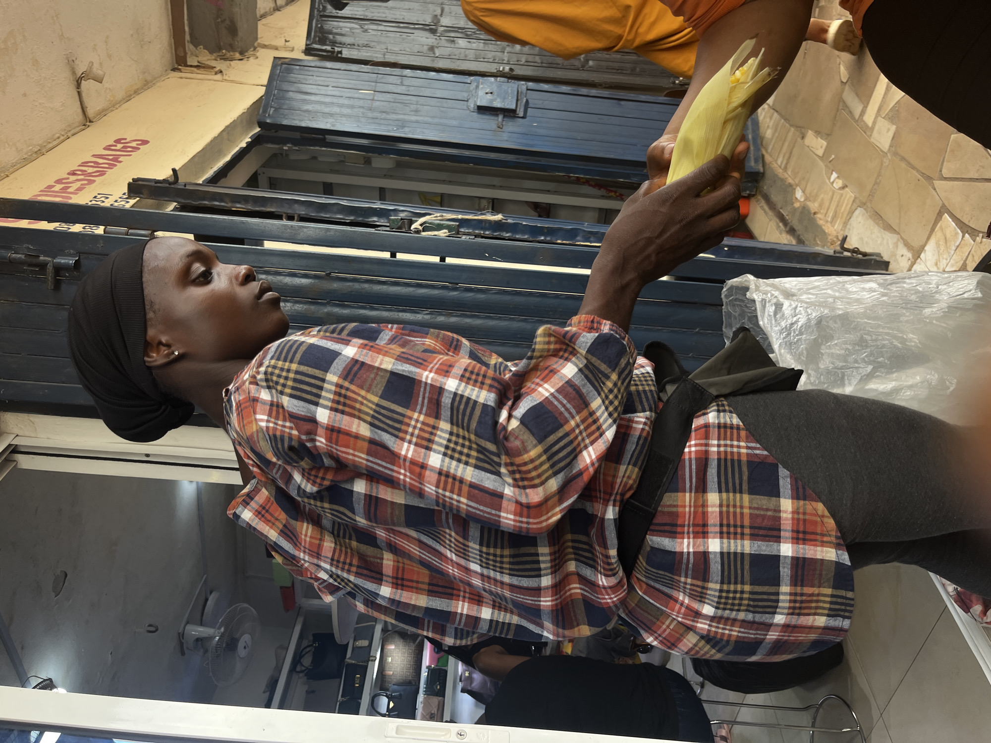 In an outdoor market stall in Jos, Nigeria, a person wearing a black head covering and a plaid long-sleeved shirt, is handing a corn on the cob to another person whose arm is visible. The stall appears to be selling bags, as indicated by a sign reading "DES&BAGS." The ground is paved with interlocking bricks, and the background shows a blue metal shutter and a storefront with shelves stocked with bags. A white plastic bag is also present.