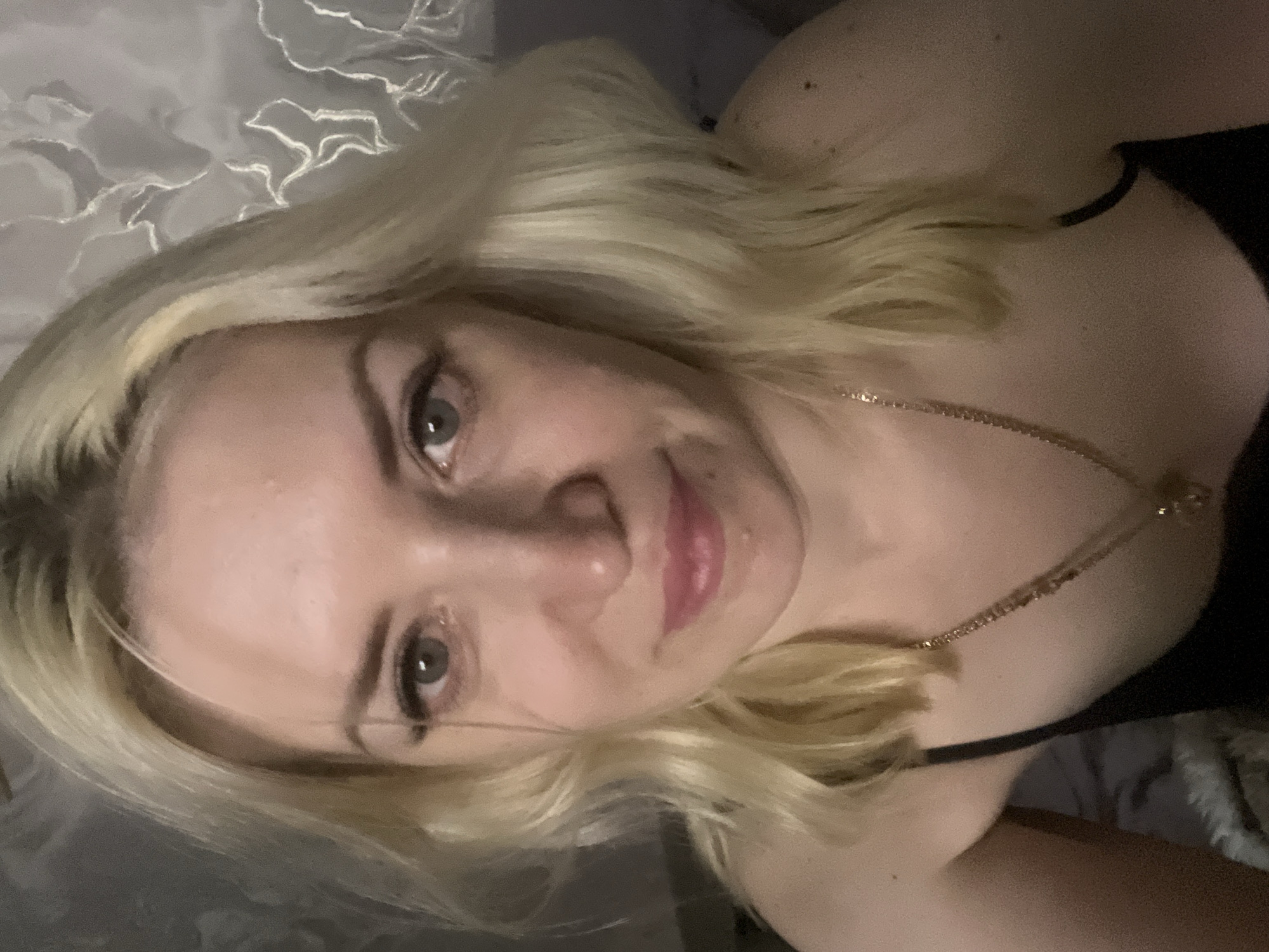 A blonde woman with light-colored eyes and pink lipstick is shown in a close-up selfie. She is wearing a black tank top and a gold necklace. The background appears to be a wall with a decorative pattern. The lighting is dim. The image does not contain any information that allows for the determination of its location in the city of Tyumen, Russia, or any other specific location.