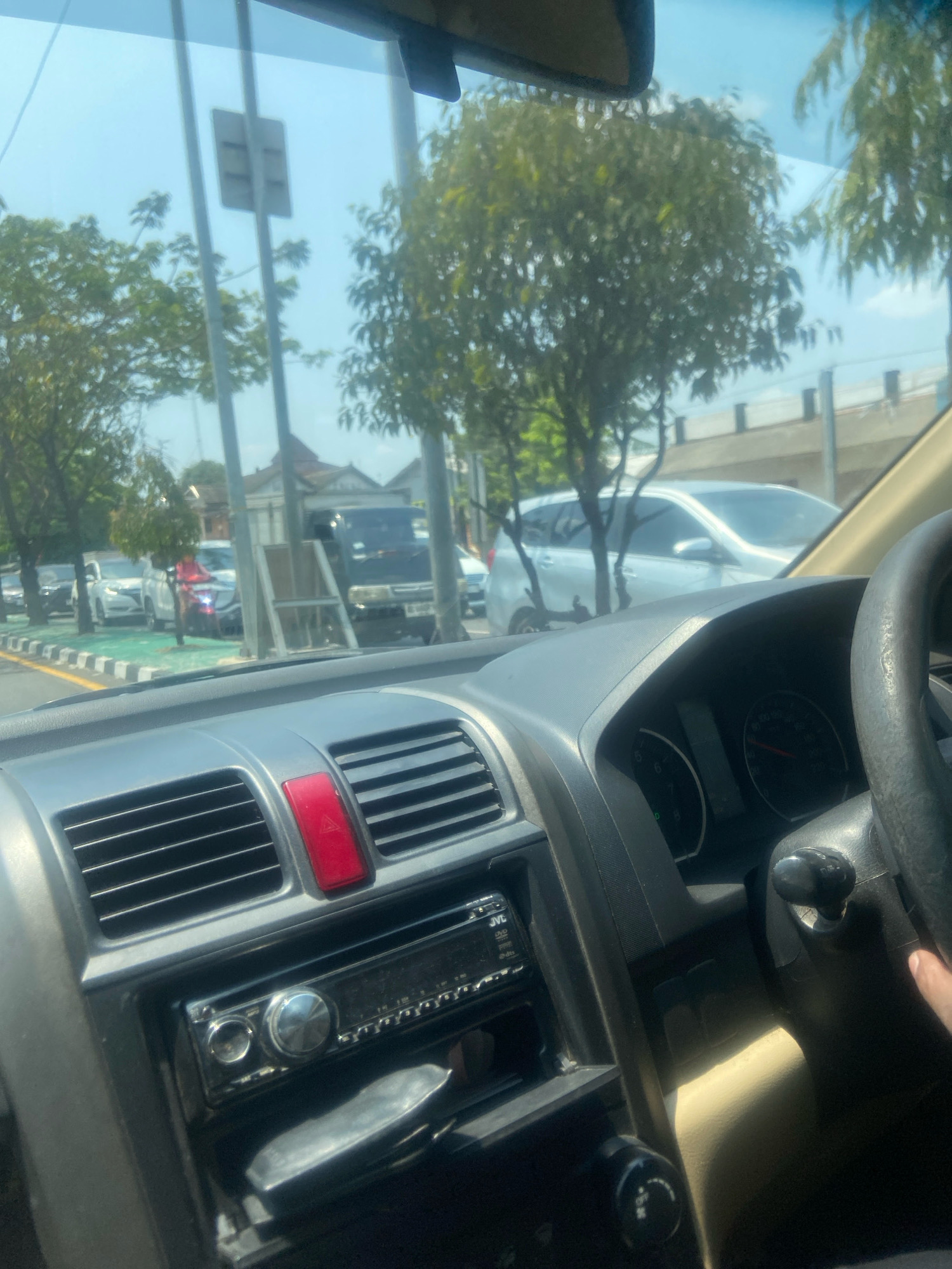 The image is taken from inside a car, looking forward and slightly to the right. The dashboard is visible in the foreground, featuring an air vent with a red hazard light button above it, and a car stereo. Beyond the windshield, a street scene unfolds under a bright sky. Trees line the roadside, with some buildings visible in the background. Several vehicles are on the road, including cars, a truck, and a motorcycle with a rider wearing a pink jacket. The overall impression is of a sunny day in a moderately busy urban or suburban area. The scene is likely captured in Ngaglik, Indonesia, given the context provided.
