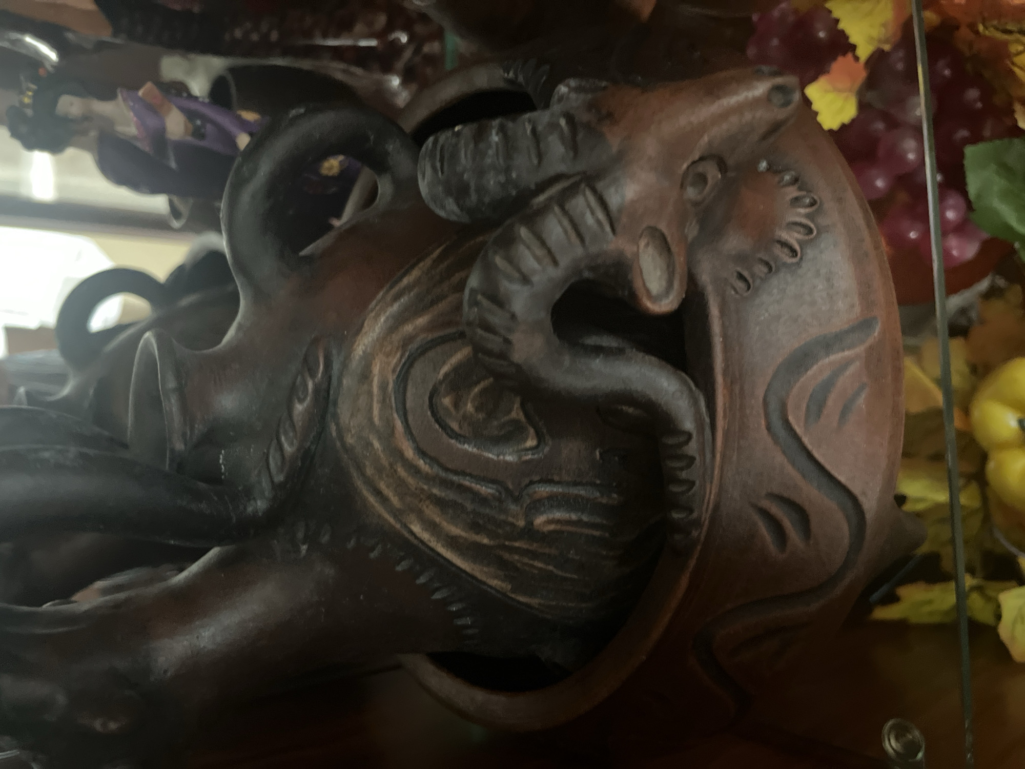 A close-up, medium shot shows a dark brown, ceramic incense burner with a detailed, stylized goat head as its focal point. The goat's head, featuring prominent horns and carved facial features, emerges from the base of the burner. Intricate carvings, possibly representing flowing lines or abstract patterns, are visible on the surface of the burner. In the background, a blurred image of a traditional Japanese geisha doll in a purple kimono can be seen, along with decorative faux grapes and leaves in shades of red and yellow. The lighting is dim, creating shadows that highlight the textures and shapes of the objects. The scene is indoors, likely on a shelf or within a display cabinet, with a reflective surface suggesting a mirrored background or glass enclosure. The exact location within Vladimirovka, Russia is not discernible from the visual information provided.