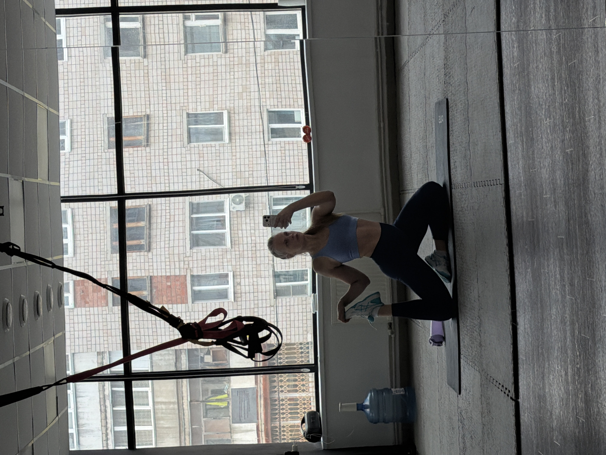 A woman is taking a selfie in what appears to be a gym with a large window showing an apartment building outside. The woman is wearing a blue sports bra and black leggings and kneeling on a yoga mat. She is holding a smartphone. There are TRX straps hanging from the ceiling. A large water bottle is on the floor near the window. The apartment building is visible through the large window and is a typical multi-story building with numerous windows. The city is Izhevsk, Russia.