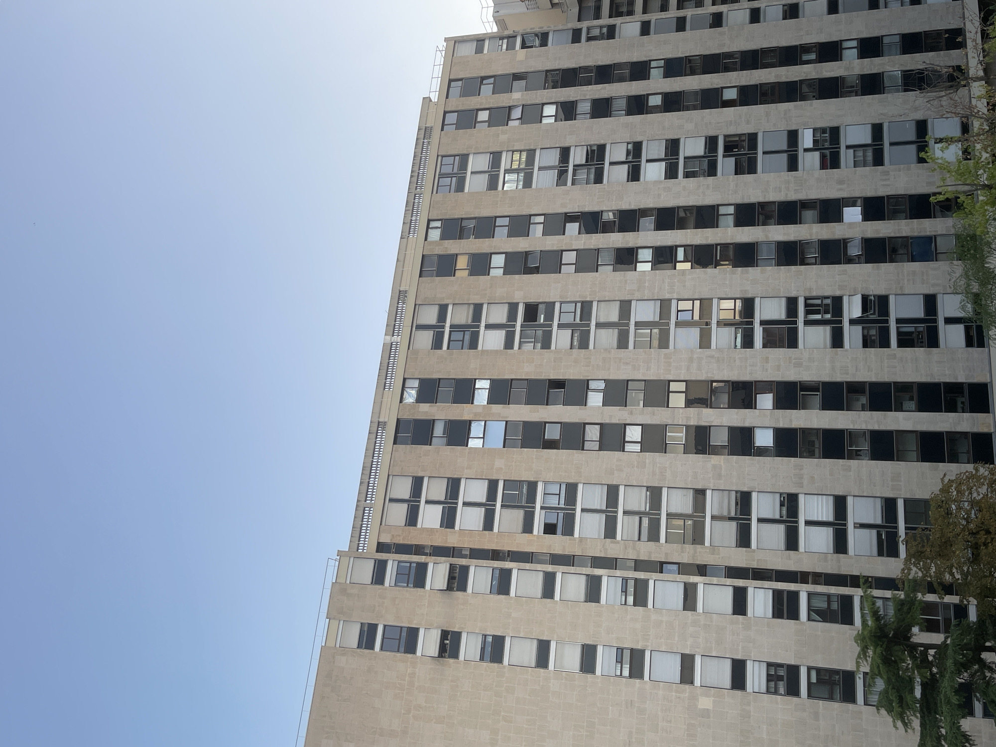 The image shows a tall, modern apartment building with a beige facade and numerous windows. The building appears to be located in a city, with a clear blue sky above. There are trees visible in the foreground, obscuring the lower part of the building. The architecture suggests a residential or office building. The windows are a mix of clear and opaque, with some blinds or curtains visible. Some windows are open, suggesting occupancy. The overall impression is one of urban development and residential living.