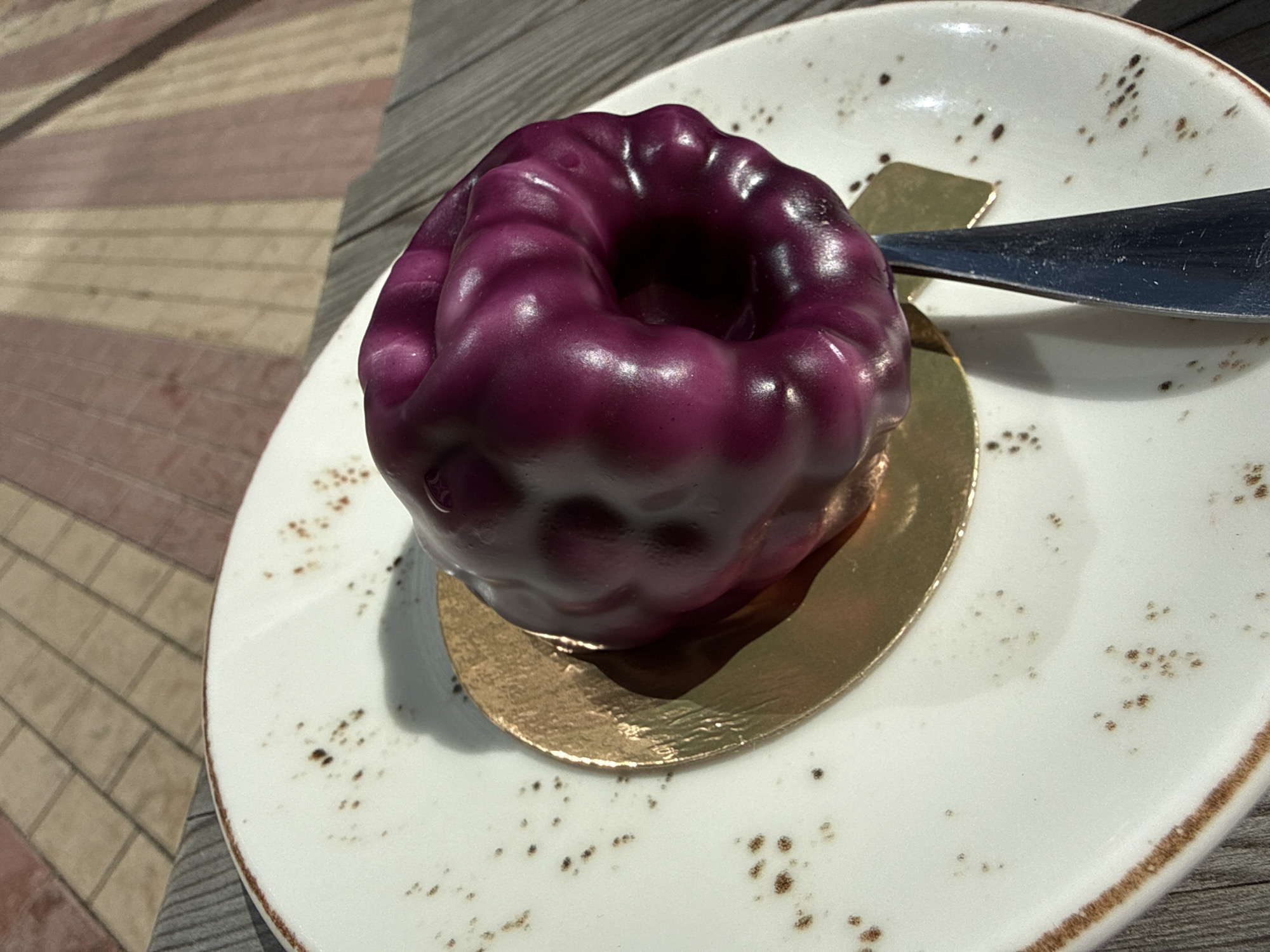 A single, small, bundt-shaped pastry with a glossy, deep purple glaze sits on a white, speckled plate. The pastry is placed on a gold-colored circular disc. A metal knife, with a dark, slightly textured handle and a shiny blade, rests on the plate to the right of the pastry. The plate is on a wooden surface, which appears to be part of a table or bench. In the background, a tiled walkway with alternating brown and tan rectangular tiles is visible, illuminated by sunlight casting shadows. The setting is outdoors, possibly a park or cafe area, in Moscow, Russia.