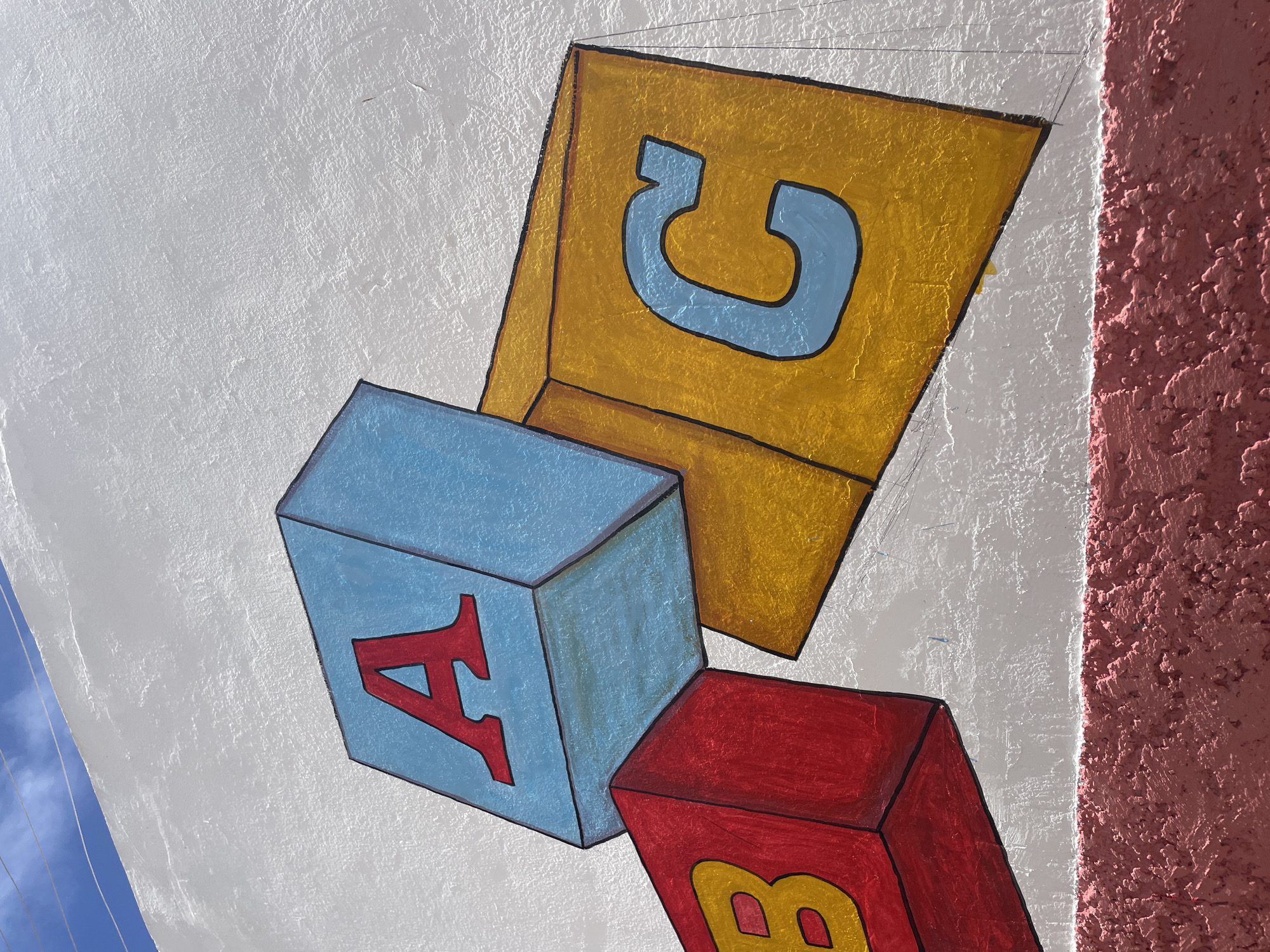The image shows a section of a white, textured exterior wall in El Valle del Espíritu Santo, Venezuela. Painted on the wall are three large, colorful toy blocks. The block on the left is red with a yellow letter 'B' on its visible side. The block in the center is light blue with a red letter 'A' on its visible side. The block on the right is yellow with a light blue letter 'C' on its visible side. The blocks are depicted in a way that suggests they are stacked or leaning against each other. The background above the wall shows a clear blue sky with thin, horizontal power lines. Below the painted blocks, there is a band of textured, pinkish-red material, likely the base of the wall or a decorative border. No people are present in the image. The scene appears to be outdoors, possibly on the side of a building.