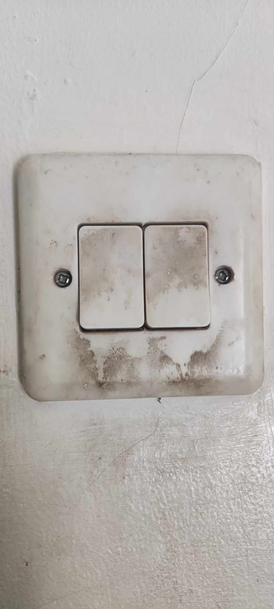A close-up, eye-level, vertical shot shows a dirty, off-white double light switch mounted on a cracked, off-white wall. The switch plate is rectangular with rounded corners and shows significant grime and dirt accumulation, particularly around the bottom edge and on the two rectangular rocker switches. Each switch has visible stains that appear to be smudges or possibly burn marks. Two silver screws are visible, one on the left side of the switch plate and one on the right, holding it to the wall. The wall itself has a textured surface and displays a prominent vertical crack extending from the top edge of the frame down past the switch. The overall impression is one of disuse and neglect. The image was taken in Gebang, Indonesia.