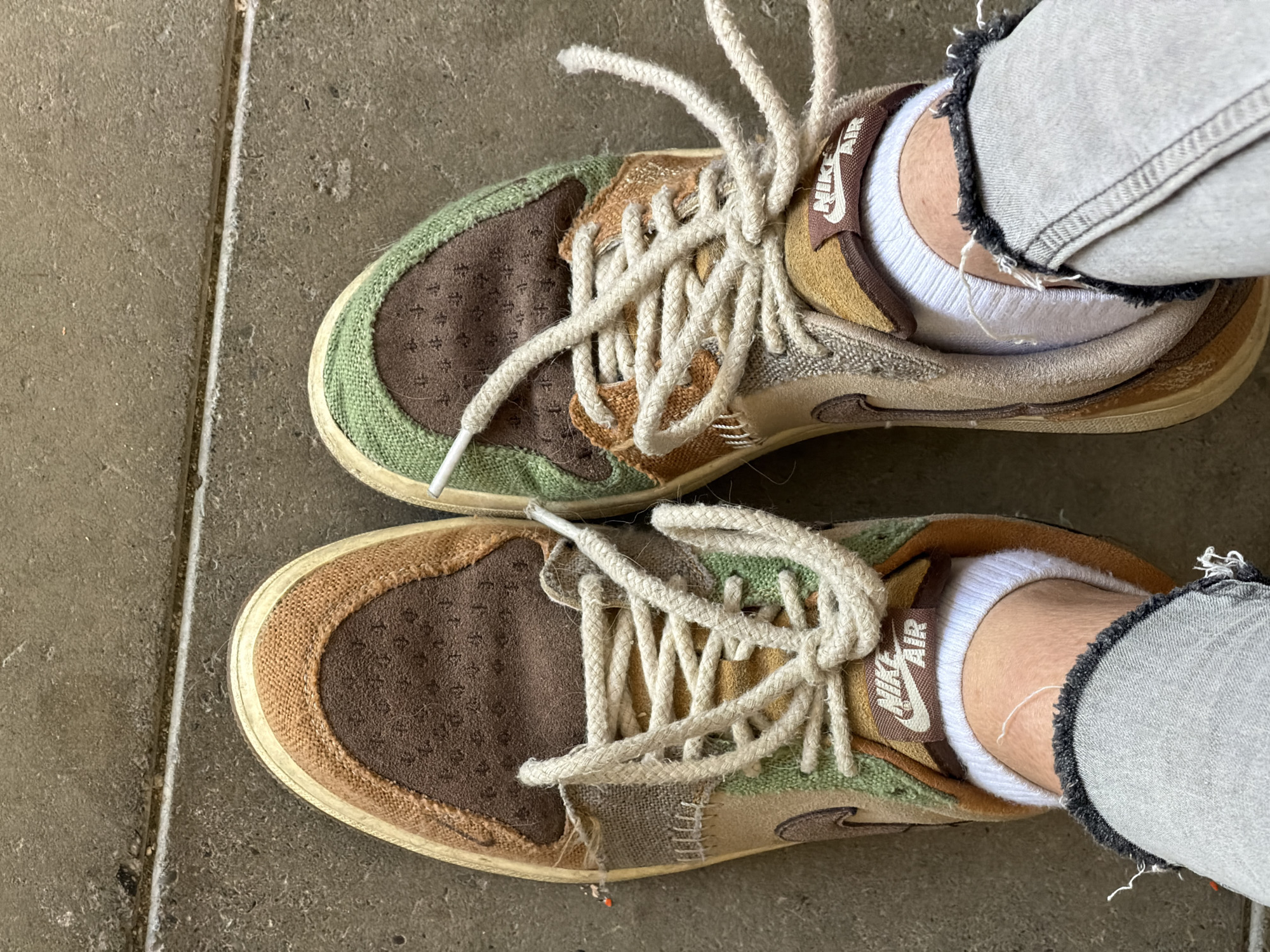 An overhead, full-frame shot captures a person's feet and lower legs, adorned with distinctive sneakers and distressed jeans, resting on a concrete surface. The scene is set outdoors on what appears to be a paved area, likely a sidewalk, characterized by a textured, light gray concrete ground with a prominent straight seam or crack running vertically through the middle of the frame.
The main subjects are a pair of sneakers featuring a multicolored, patchwork design. The shoes display an intricate mix of earthy tones including various shades of brown, green, beige, and tan, crafted from different materials such as what appears to be suede, woven fabric, and canvas. Thick, off-white, rope-like laces are loosely tied on both shoes. A brown tag with the text "NIKE AIR" in white lettering is visible on the tongue of each sneaker. The soles are a light, slightly cream-colored hue, showing subtle signs of wear.
The person is wearing light gray jeans, notably distressed with rips and frayed edges around the ankles. White crew or ankle socks are visible between the top of the shoes and the frayed denim. The lighting is bright and even, suggesting it is daytime. While the location context is Tbilisi, Georgia, no specific visual cues within the image confirm this, only general urban elements like the concrete ground.