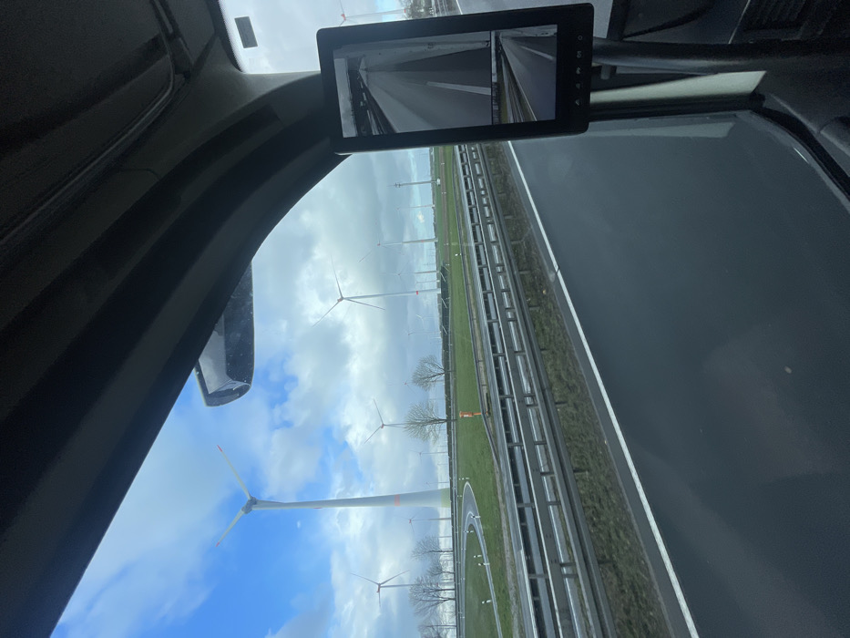 The image depicts an exterior highway scene observed from inside a vehicle, likely a truck, in Marsberg, Germany. The vehicle's dark gray interior A-pillar and roof lining are visible at the top and right. A vertical digital display acting as a rearview mirror is mounted on the interior right, showing a split-screen view of a wet road surface. The primary view through the windshield shows a multi-lane highway with dark, wet asphalt, featuring a white solid lane line on the right. To the left, a metal guardrail system separates the main highway from a green grassy median or embankment. Beyond this, a curved asphalt access road or ramp, marked with white dashed lines and small white poles, is visible, surrounded by green grass. The background features a large wind farm with numerous white wind turbines with three blades extending across the landscape. The closest large turbine on the left displays a red band near the base of its tower. Scattered bare deciduous trees are visible in the grassy areas. The sky is partially cloudy with patches of blue. No people, animals, or explicit actions or interactions are visible.