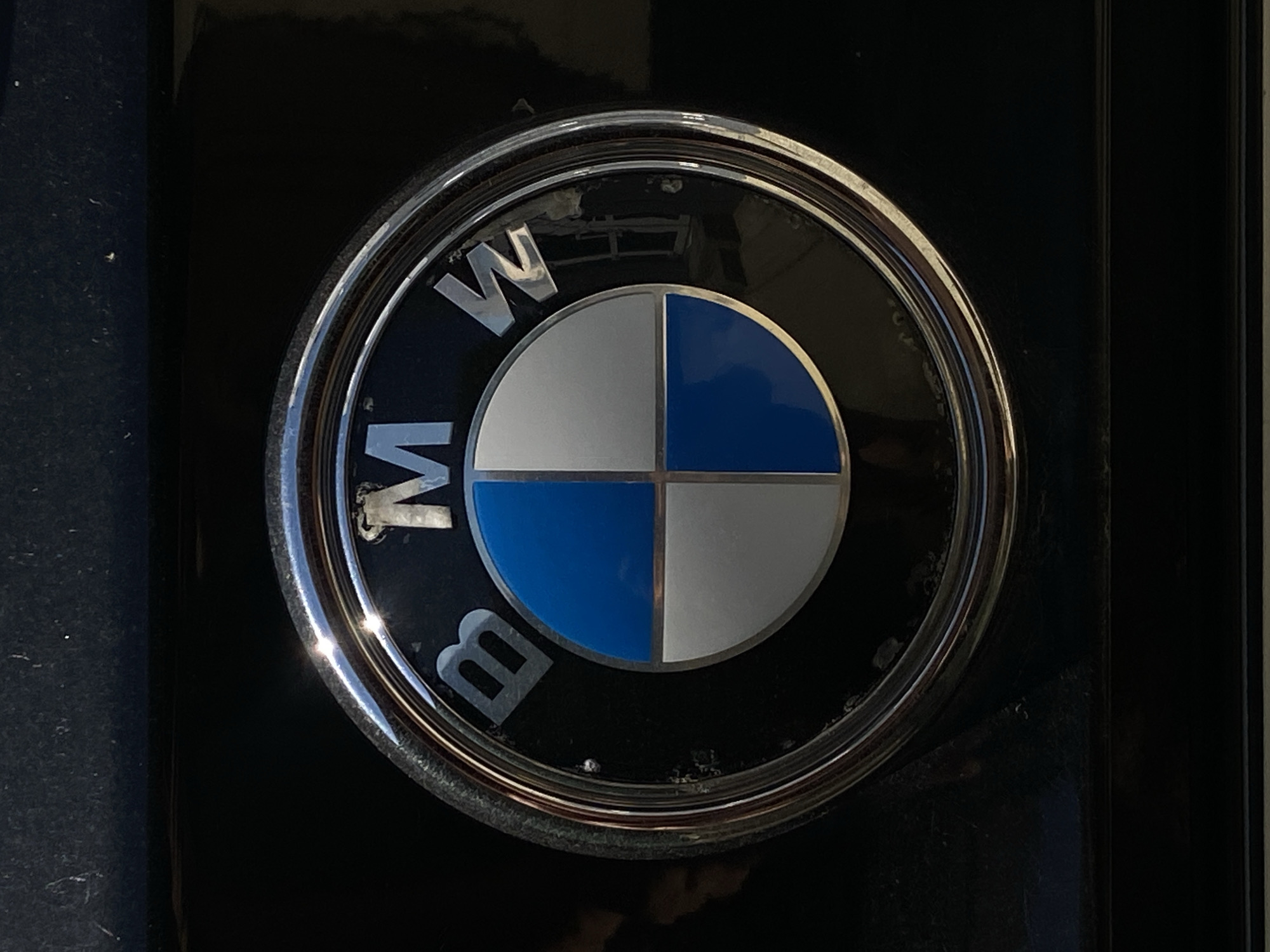 A close-up shot of a BMW car logo affixed to a black surface. The circular logo has a chrome outer ring with the letters "BMW" in capital letters embedded within. The central part of the logo is divided into four quadrants, alternating between blue and silver/white. The logo shows signs of wear and age. The surrounding surface appears to be a dark, possibly painted panel. The location in the city of Vysokoye, Russia is not discernible from the image itself.