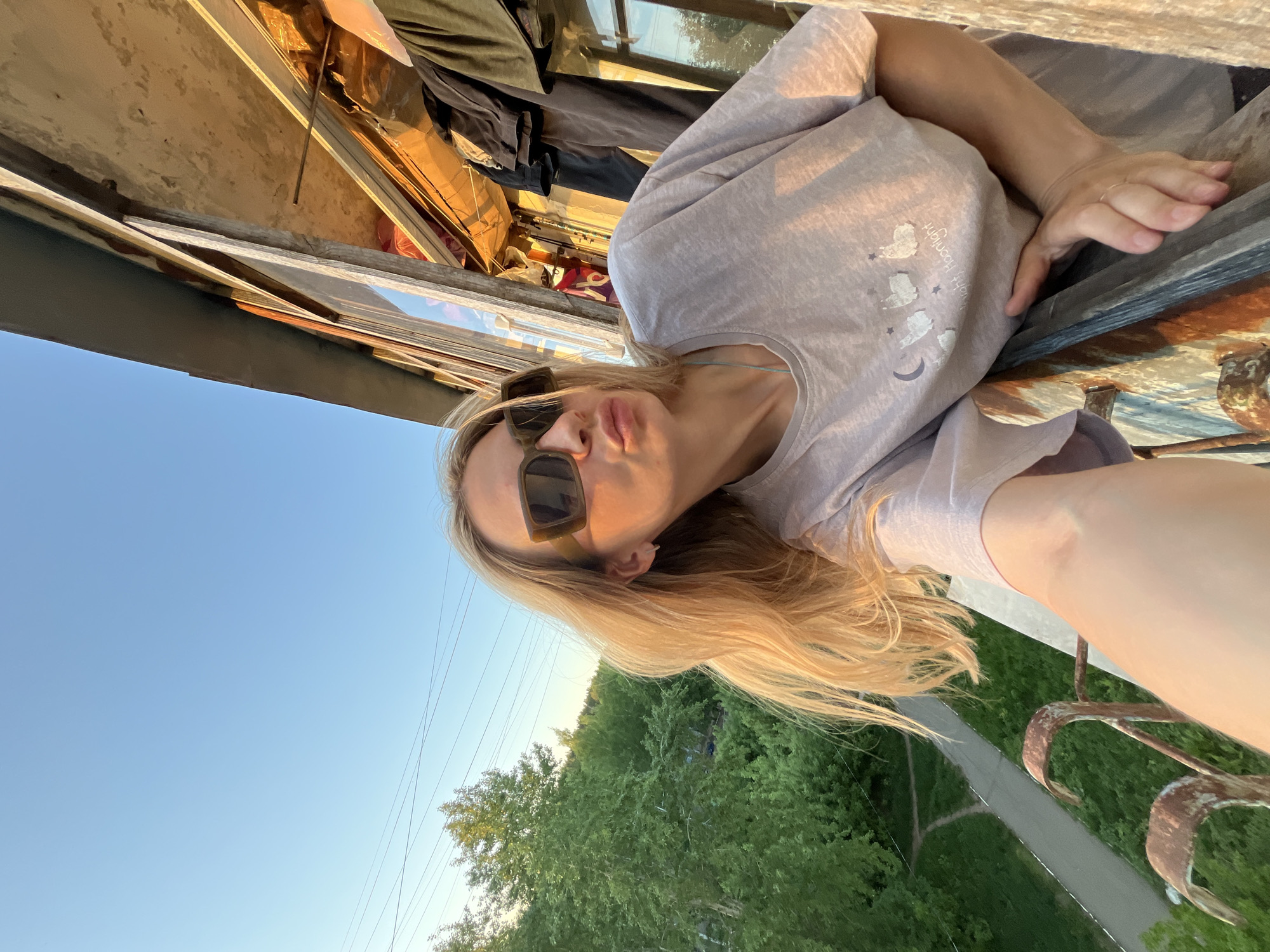 A woman with blonde hair and sunglasses is taking a selfie outdoors. She is wearing a light-colored t-shirt with a graphic design. Behind her, there is a view of a clear blue sky, green trees, and a street. The location appears to be an elevated position, possibly a balcony or rooftop. There is construction material in the background and some corroded metal railing. There is not enough detail to confirm the city as Chaikovsky.