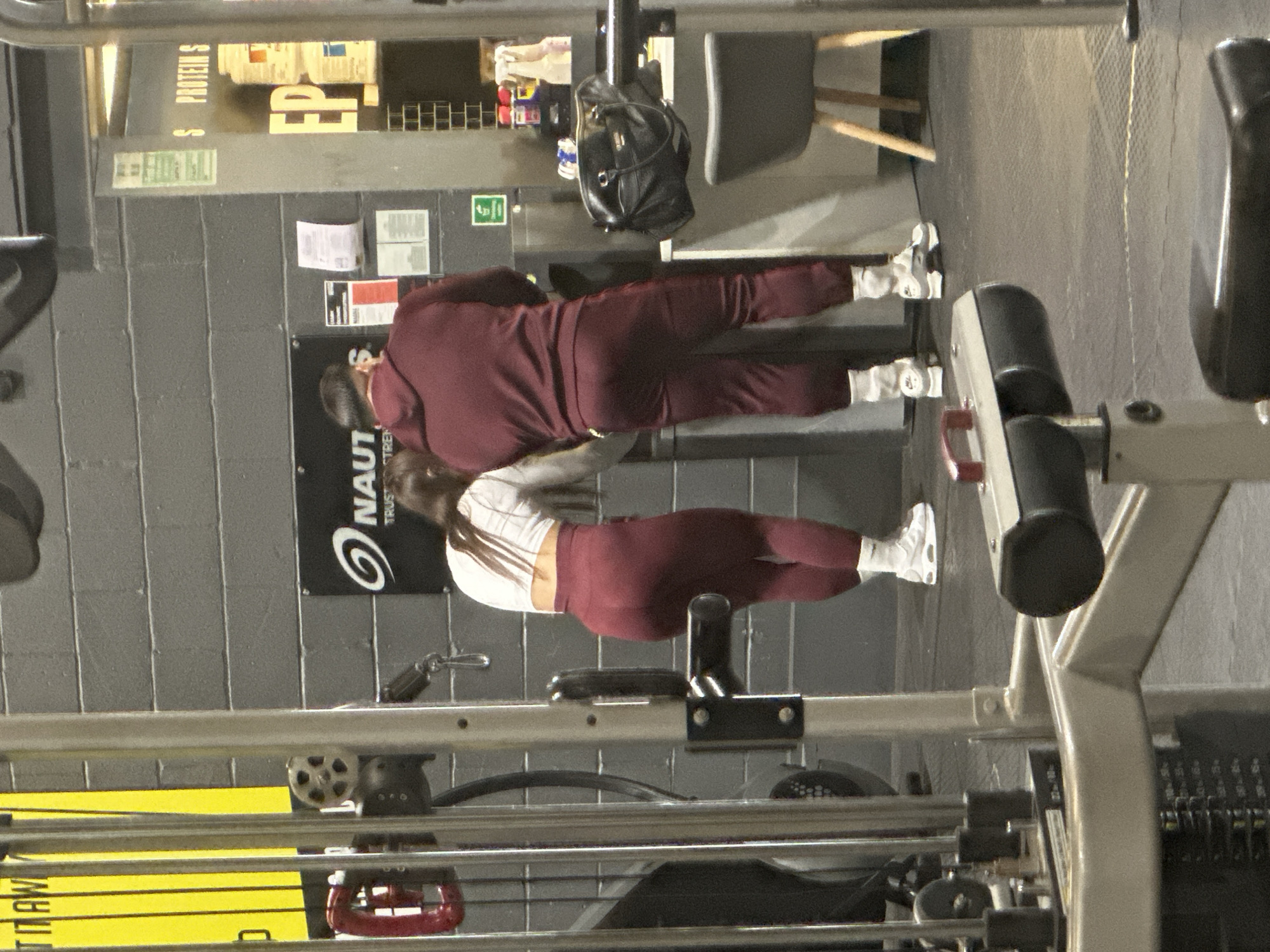 A sideways indoor shot shows two people exercising at a gym in Tonbridge and Malling, United Kingdom. The male is wearing a maroon tracksuit, while the female is wearing a white top and maroon leggings. They both have white sneakers on. The man is positioned above the woman and facing away from the camera, while the woman is below him and facing the camera. Various exercise machines are in the foreground and background.