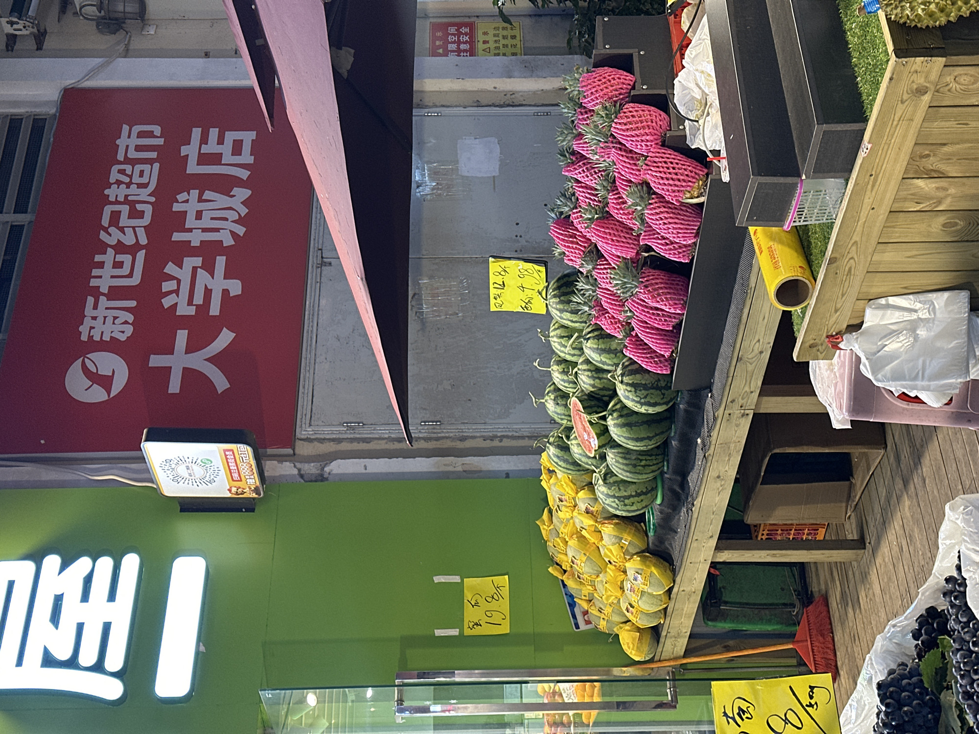 The image captures a fruit stand in Chongqing High-tech Zone, China, showcasing various fruits, including pineapples wrapped in pink netting, watermelons, yellow-wrapped cantaloupes, and grapes. The fruits are arranged on a wooden shelving unit. A sign in Chinese advertises the prices of the fruits. In the background, there's a sign for a "New Century Supermarket" (新世纪超市), and another sign displaying a QR code. The overall setting suggests an outdoor fruit market or a produce section of a supermarket.