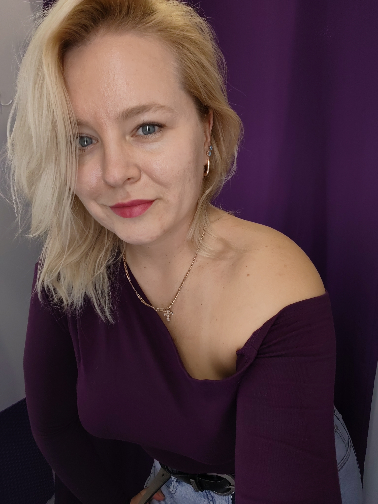 A head and shoulders shot shows a Caucasian woman with blonde hair, blue eyes, and pink lipstick. She is wearing a plum-colored off-the-shoulder top and a gold necklace with a cross pendant. The background is a blurred purple curtain. The image is a selfie, as indicated by the angle and the woman's gaze into the camera. There is no identifiable information about her location in Izhevsk, Russia, or any other specific location.
