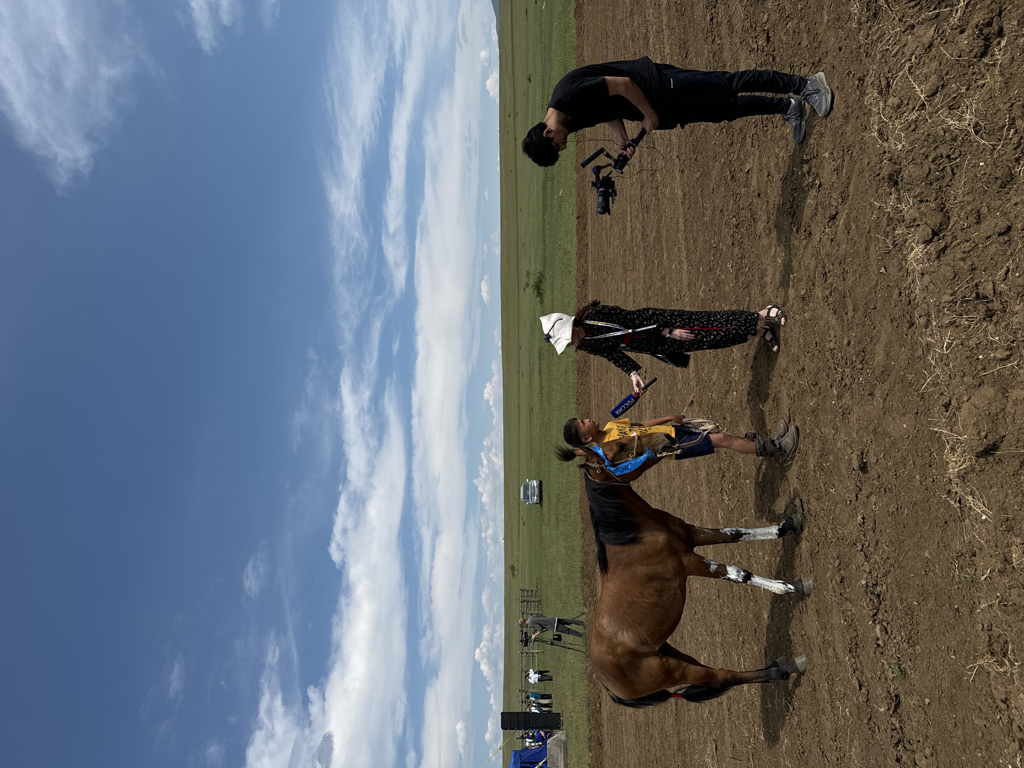 In the city of Orongoi, Russia, on a clear day with scattered clouds, a young boy is being interviewed next to a brown horse. The boy, wearing a yellow shirt and blue shorts, stands to the right of the horse, holding its reins. A woman in a black dress and white hat is interviewing him, holding a microphone. To the right of the woman, a man is filming the scene with a professional camera. In the background, a vast green field stretches to the horizon, with a few trees and a white car visible. A blue tent and a group of people are also present in the distance. The ground in the foreground is bare earth.