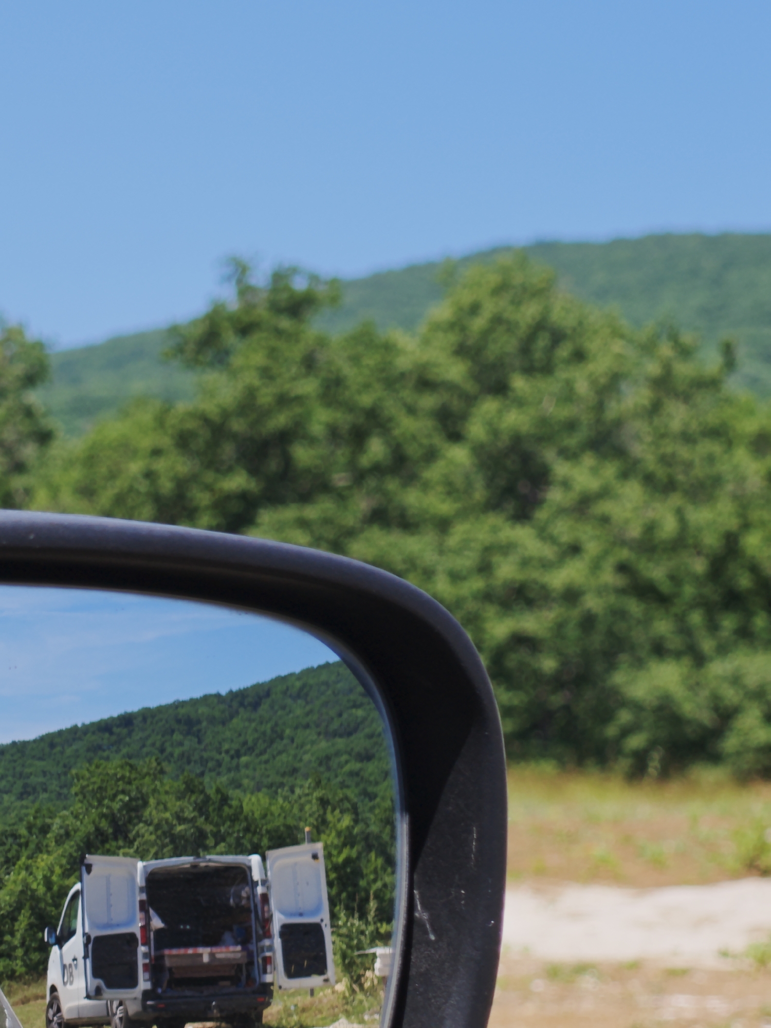 In Izgrev, Bulgaria, a white van with its rear doors open is parked on a dirt road. The van has the letters "DB" on its side. The scene is reflected in the side mirror of a vehicle, showing a landscape of green trees and hills under a clear blue sky. The reflection also includes some dry grass and brush in the foreground.