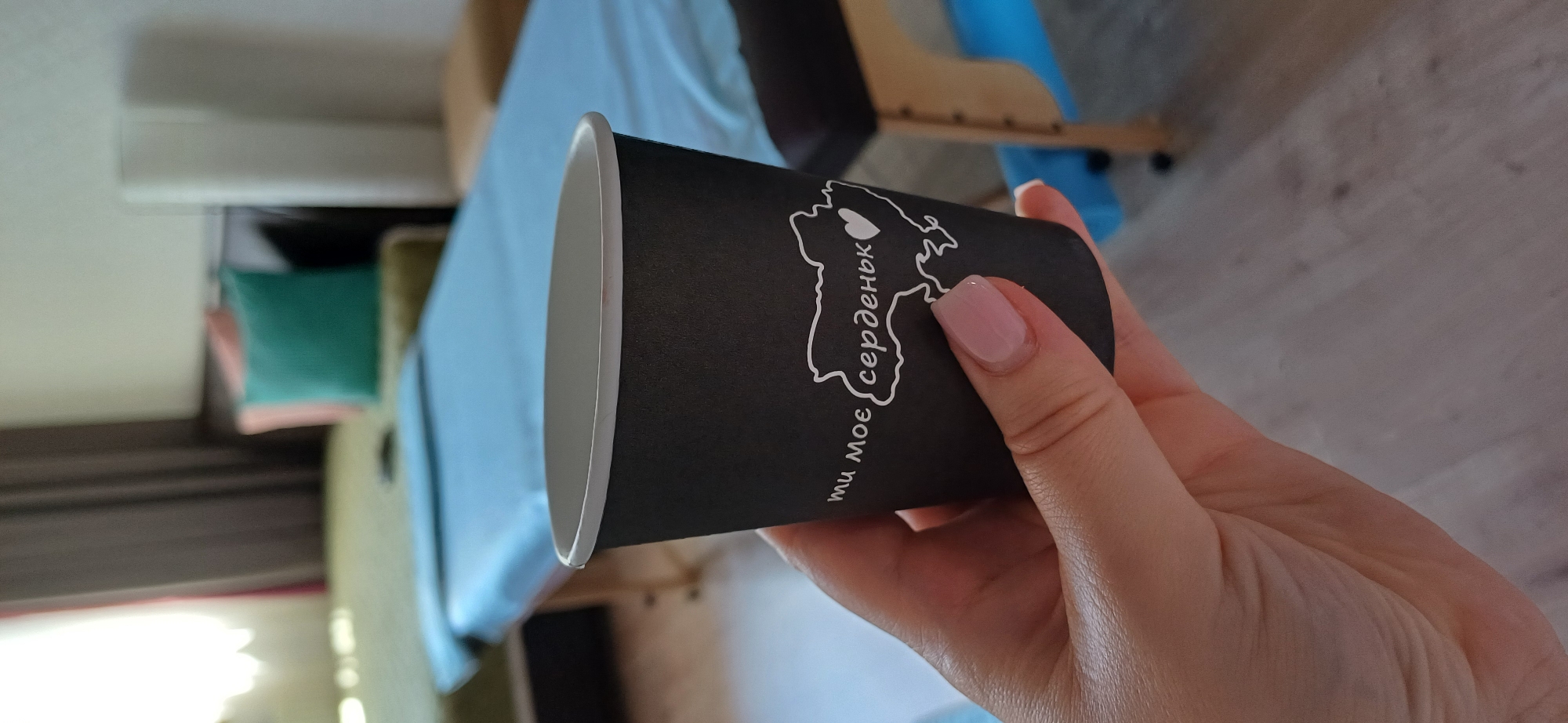 A black paper cup with white text and a white outline of Ukraine with a heart in the center is held in a woman's hand. The text on the cup reads "ти моє серденьк". The background of the image is blurred, but appears to be a room with a massage table covered in a light blue sheet. Sunlight is streaming in from a window on the left side of the frame. The location is not specified in the image.
