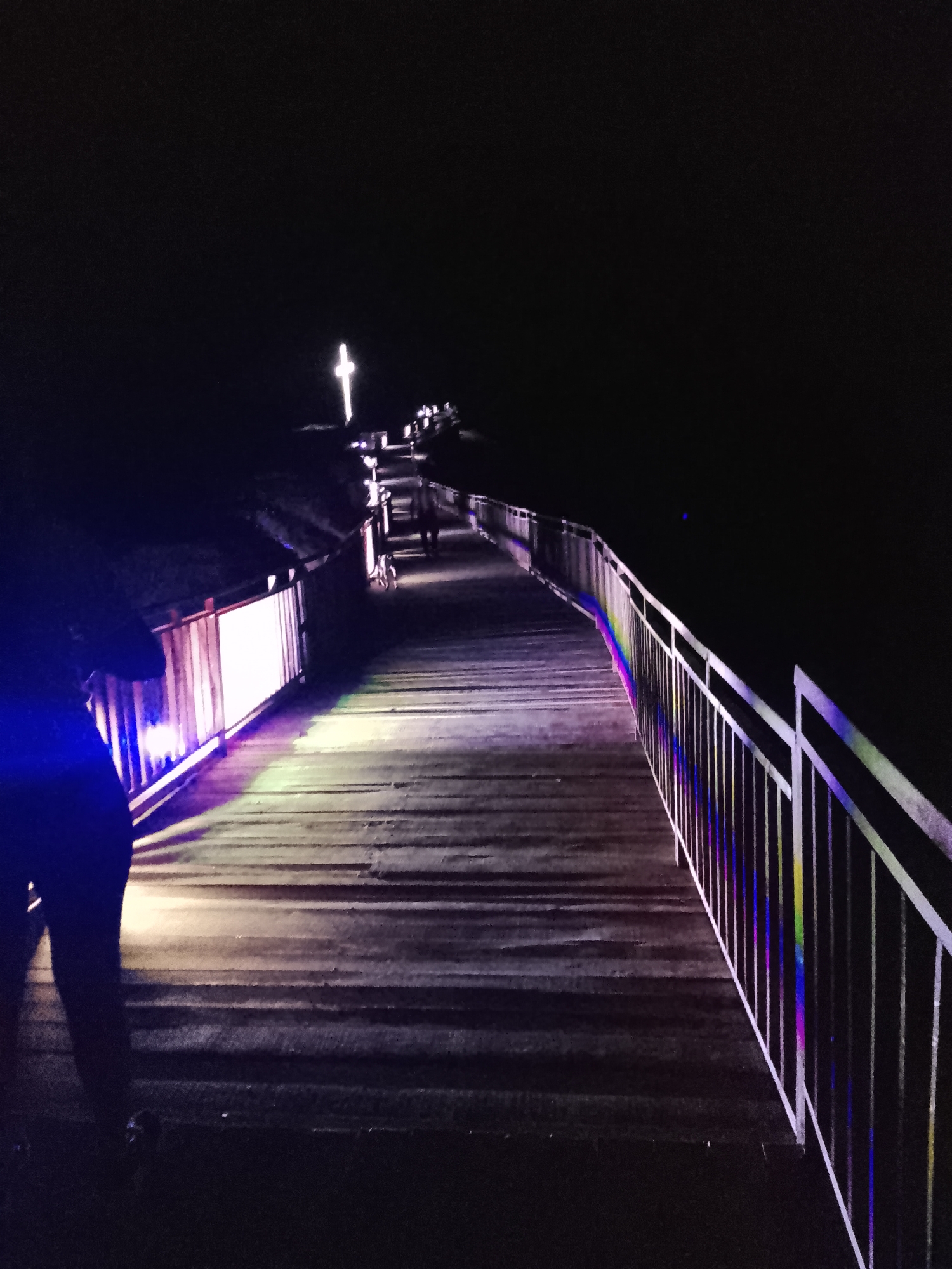 The image shows a wooden boardwalk at night, illuminated by artificial lights. The boardwalk extends into the distance and is lined with a white metal railing that reflects colorful light. In the background, a large illuminated cross is visible, suggesting a religious or memorial site. There are a few silhouetted figures on the boardwalk, some walking. The overall atmosphere is dark and somewhat mysterious. The location is identified as the "Provadia, Bulgaria" memorial.