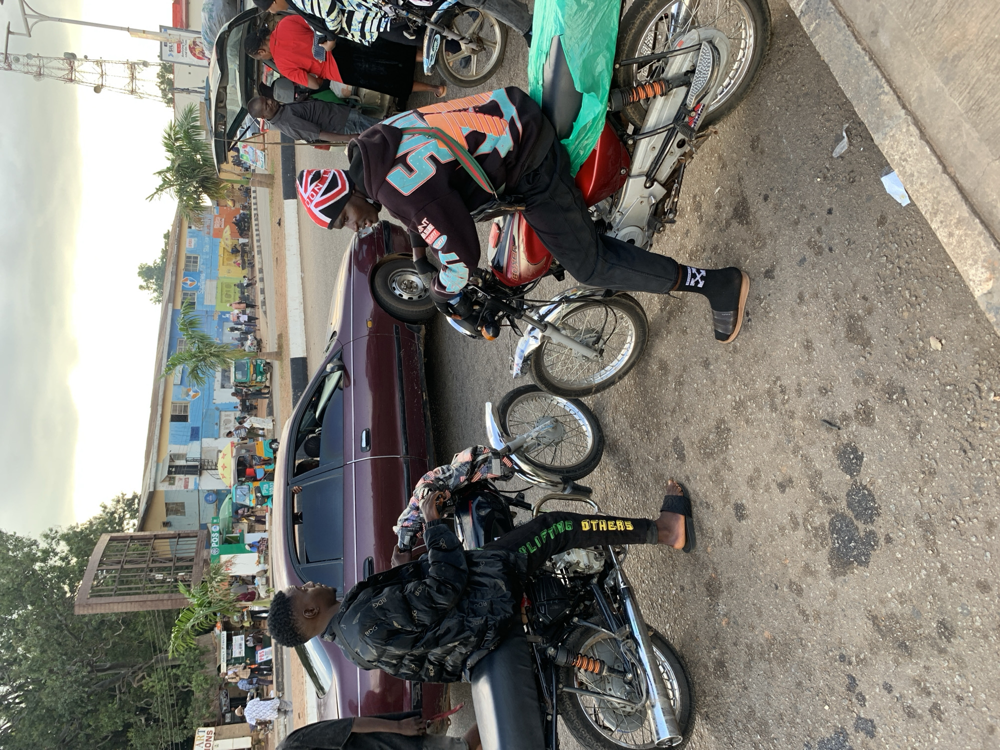 In Jos, Nigeria, two young men are seated on motorcycles. The man on the left wears a black puffer jacket and black pants with green lettering that says "LIFTING OTHERS". He has black slides on his feet. The man on the right wears a black hoodie with colorful graphics and a red, white, and blue beanie. He has black socks and brown slides. Both men are looking at each other. The motorcycles are parked on a paved road with some oil stains and debris. In the background, there is a maroon sedan, other vehicles, buildings, and trees, suggesting a busy street scene. The sky is overcast.
