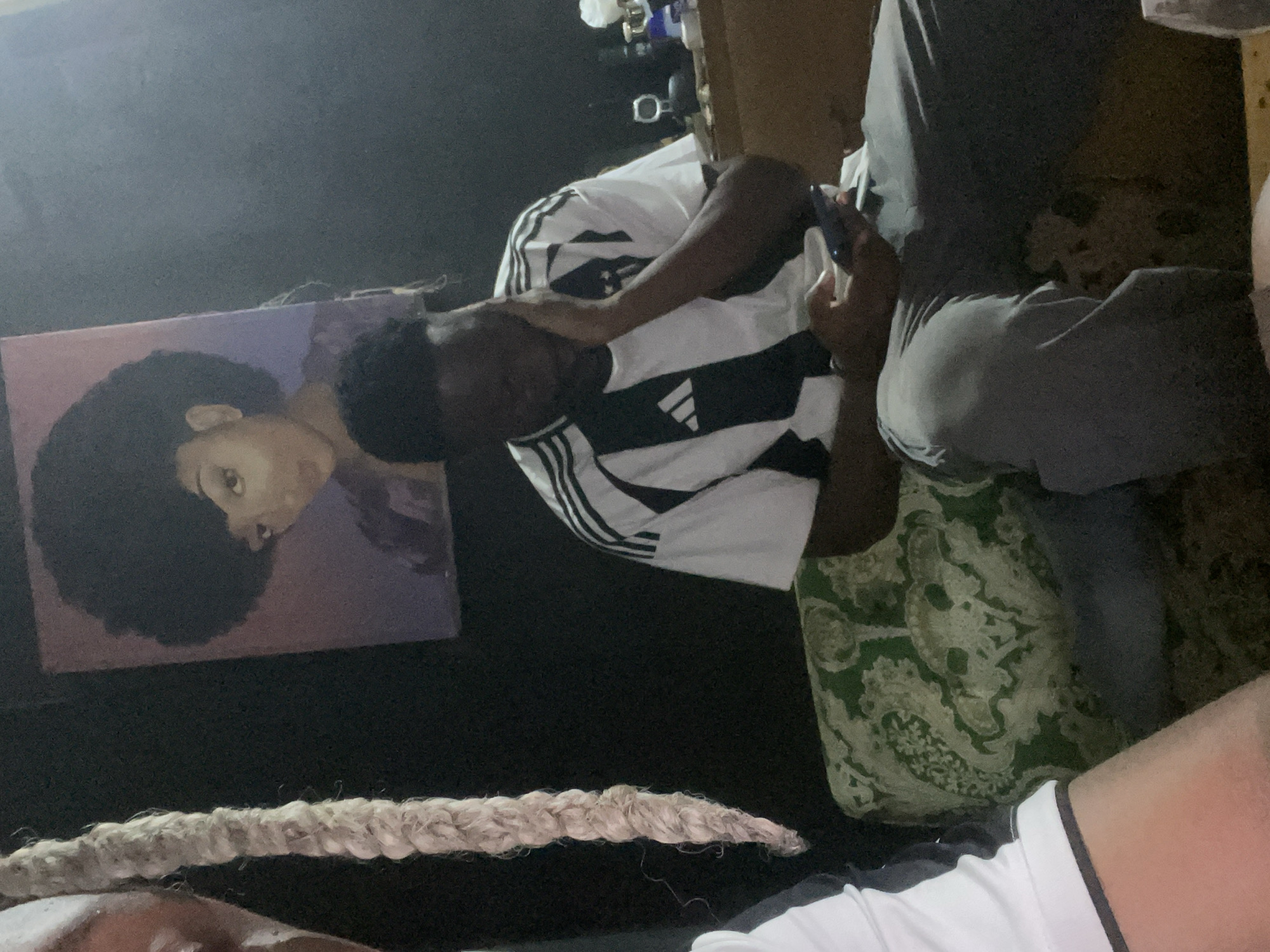 In an indoor setting, possibly in Jos, Nigeria, two individuals are present. On the left, a person with long, light-colored dreadlocks is partially visible, with their arm and shoulder in the foreground, wearing a white shirt with dark trim. To the right, a young Black male wearing a black and white striped athletic shirt is seated. He is holding a dark-colored smartphone in his right hand and resting his left hand on his face, appearing to be looking down at the phone. He is seated on a patterned cushion with green and white designs. Behind him, a framed painting of a woman with an afro hairstyle hangs on a dark wall. To the far right, a wooden surface with various items, including bottles and a cylindrical object, is visible. The lighting in the room appears dim, with some light sources casting shadows.