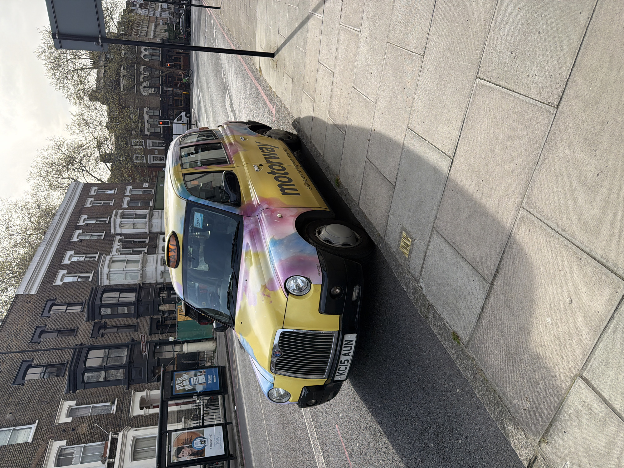 A London taxi, distinguished by its colorful paint scheme featuring the word "motorway" and registration number KC15 AUN, is parked on a street next to a sidewalk. The taxi's signage is visible. The street and sidewalk are wet, potentially indicating recent rainfall. Adjacent to the taxi is a bus stop with advertisements for products, including "Clearspring TOFU". In the background are London townhouses of brick, some of which appear to have black painted trim, and leafless trees. A building bears the name "SADLERS WELLS".
