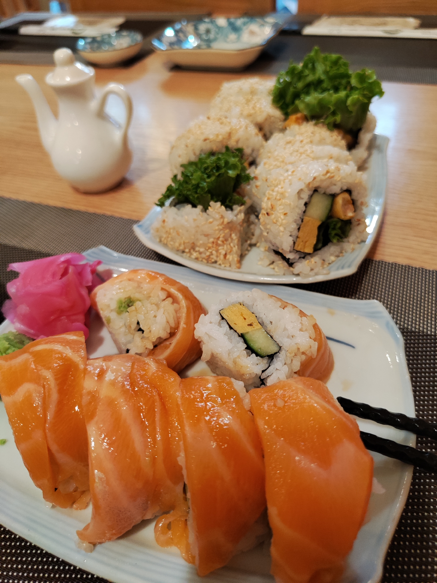 The image shows two plates of sushi on a wooden table with a placemat. The closer plate contains salmon sushi, along with pickled ginger, wasabi, and black chopsticks. The second plate holds sesame seed-covered sushi rolls filled with lettuce, cucumber, and what appears to be egg. A small white ceramic pitcher is visible in the background to the left. Additional plates are placed in the background. Based on visual appearance and style, it is likely the sushi was prepared in Da Lat, Vietnam, but there is no definitive proof.