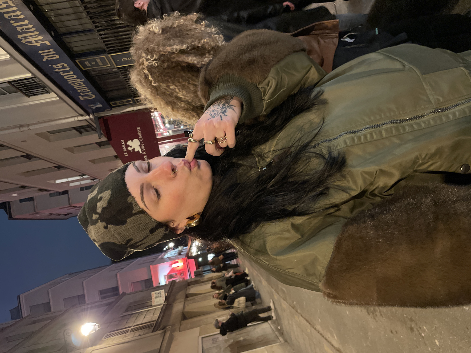 The image depicts a nighttime street scene in the Marais district of Paris, France. A female subject is positioned in the foreground, facing the camera with closed eyes and pursed lips, her left index finger touching her mouth. She wears a dark camouflage-patterned beanie, an olive green bomber jacket featuring dark brown fur trim on the collar and cuffs, and large gold-toned hoop earrings. A small silver ring is visible in her right nostril. Her left hand displays multiple gold rings on the index and middle fingers, along with a black ink floral tattoo on the back of her hand. Her hair is long, dark, and straight.
In the mid-ground, to the right of the primary subject, the top of a person's head with light brown, curly hair is partially visible. Further back and to the left, a group of several blurred individuals stands on the sidewalk. The street is lined with multi-story Parisian buildings under a dark blue night sky. A warm yellow streetlamp illuminates the left side of the scene. Visible business signs include a blue sign with gold lettering reading "Transports du Marais" and a red awning displaying "LE BARAV - BAR À VIN - CAVISTE." A "VENDRE" (For Sale) sign is also affixed to a building on the left. Red light emanates from a distant storefront further down the street.