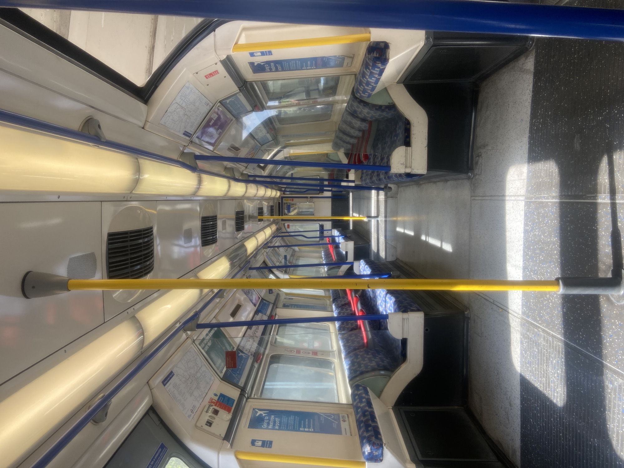 This media file captures the interior of an empty London Underground train carriage, presented rotated 90 degrees to the right. The view extends down the length of the car, showcasing rows of blue and white patterned seats on both sides. Overhead, a continuous line of bright fluorescent lights illuminates the carriage. Yellow and blue handrails are visible, running along the ceiling and descending towards the floor.
No people are present, indicating either an off-peak time, a train awaiting service, or one between stops. Strong, distinct shadows are cast across the dark grey, speckled floor from the windows, suggesting it is a sunny daytime. Various information panels and maps are affixed to the walls above the windows and seats, characteristic of public transport in London, United Kingdom.
Visible text includes multiple instances of "Going to Heathrow airport?" on prominent blue advertising panels. Other legible text snippets are "elty fare or prosecution" and "SAYS HELP," likely from warning signs or other informational posters within the train. The design of the carriage, including the seat patterns and distinctive handrails, firmly places the setting within the London Underground network.