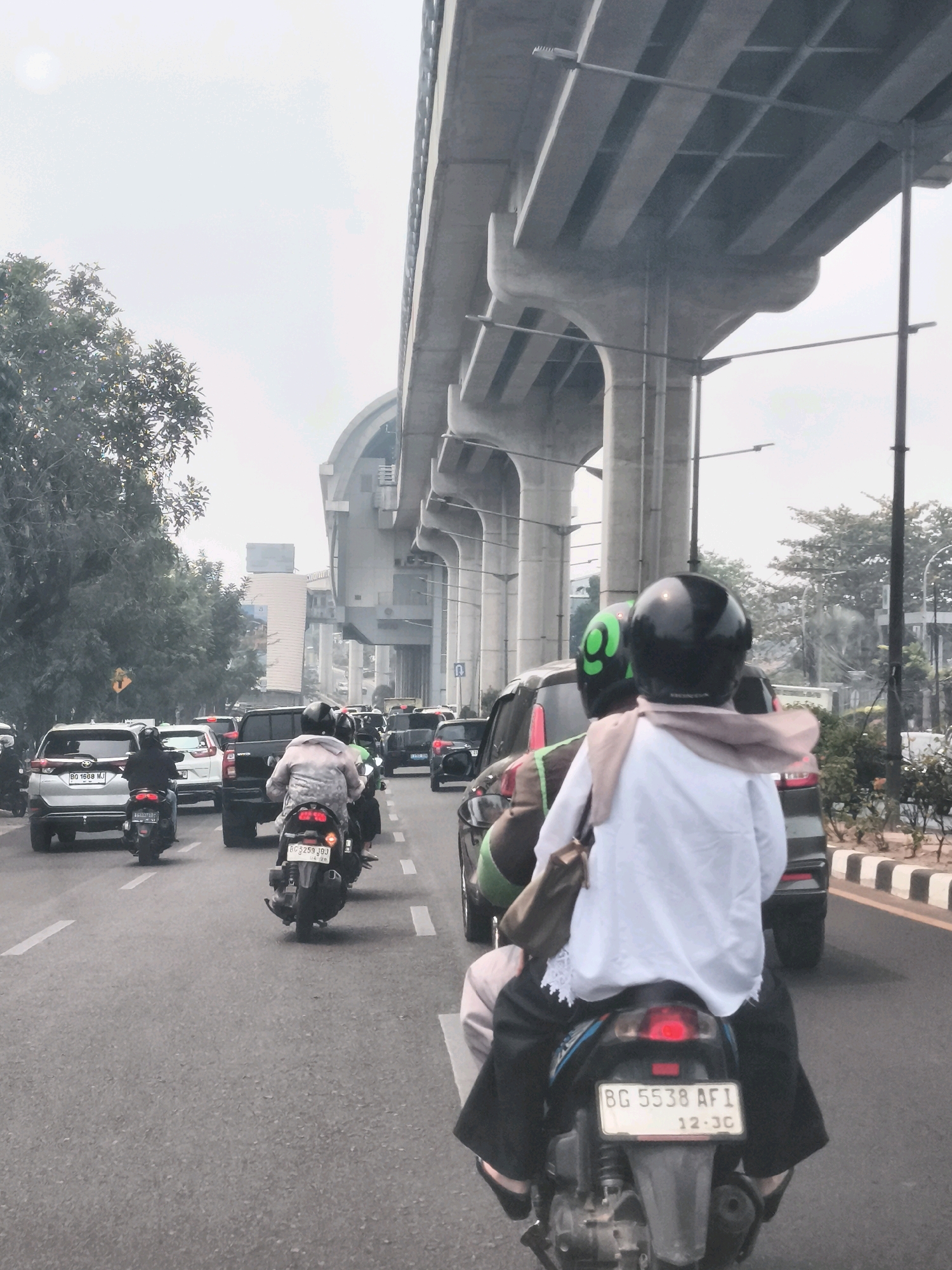 This image depicts a traffic scene on a road in Palembang, Indonesia, likely under an elevated highway structure. In the foreground, two individuals are riding on a black motorcycle, with the passenger wearing a white shirt and black pants, and a light brown scarf around their neck. The motorcycle's license plate reads "BG 5538 AFI 12-30". To the left, another motorcycle with a rider wearing a patterned jacket is visible. Further ahead, several cars and motorcycles are in traffic. The environment includes a grey sky, trees lining the road on the left, and the imposing concrete supports of an elevated structure overhead. The road surface is asphalt with white lane markings.