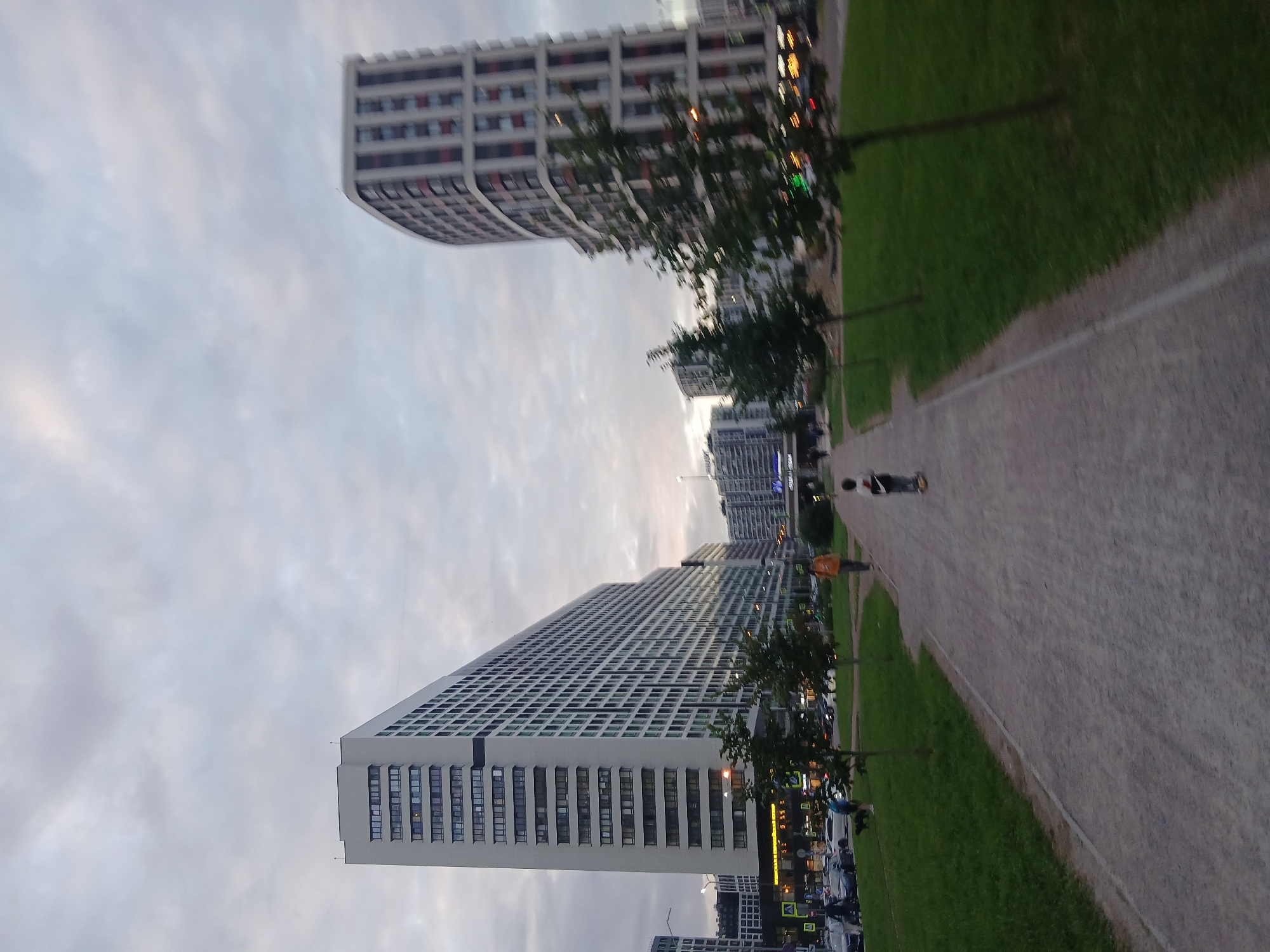 This is a ground-level, slightly low-angle, wide shot taken in Murino, Russia, looking down a park path towards large, modern apartment buildings under a cloudy sky. The path is made of light-colored gravel and is bordered on the left by a strip of grass and then a road with parked cars. On the right, the path is bordered by a grassy area with young trees. In the distance, on the left, is a very long, white, multi-story apartment building with many windows. To the right of that, further in the distance, is a curved, multi-story apartment building with red and white accents. People are visible: a child on a scooter is in the foreground on the path. Further back on the path, a person in an orange jacket is walking. On the grass to the left, a person is walking a dark-colored dog. Cars are parked along the street bordering the park. The overall impression is of a residential urban environment.