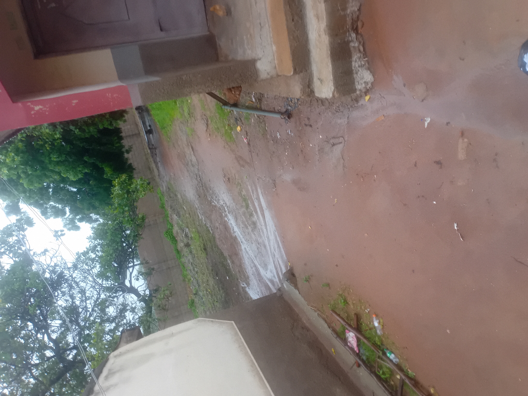 The image depicts an outdoor scene in Jauro Yinu, Nigeria, with a focus on a muddy path and the side of a building. A light-colored wall of a building is visible on the left, contrasting with the reddish-brown, wet ground. The path appears to be a dirt road, with puddles and water flow suggesting recent rain. In the background, there is a wall, green vegetation, and trees. On the right side of the image, a portion of a building with red and grey sections is present, featuring a door and some steps. Various objects are scattered around, including a pickaxe leaning against the building, some debris on the ground, and a plastic bottle. The lighting suggests an overcast or cloudy day.
