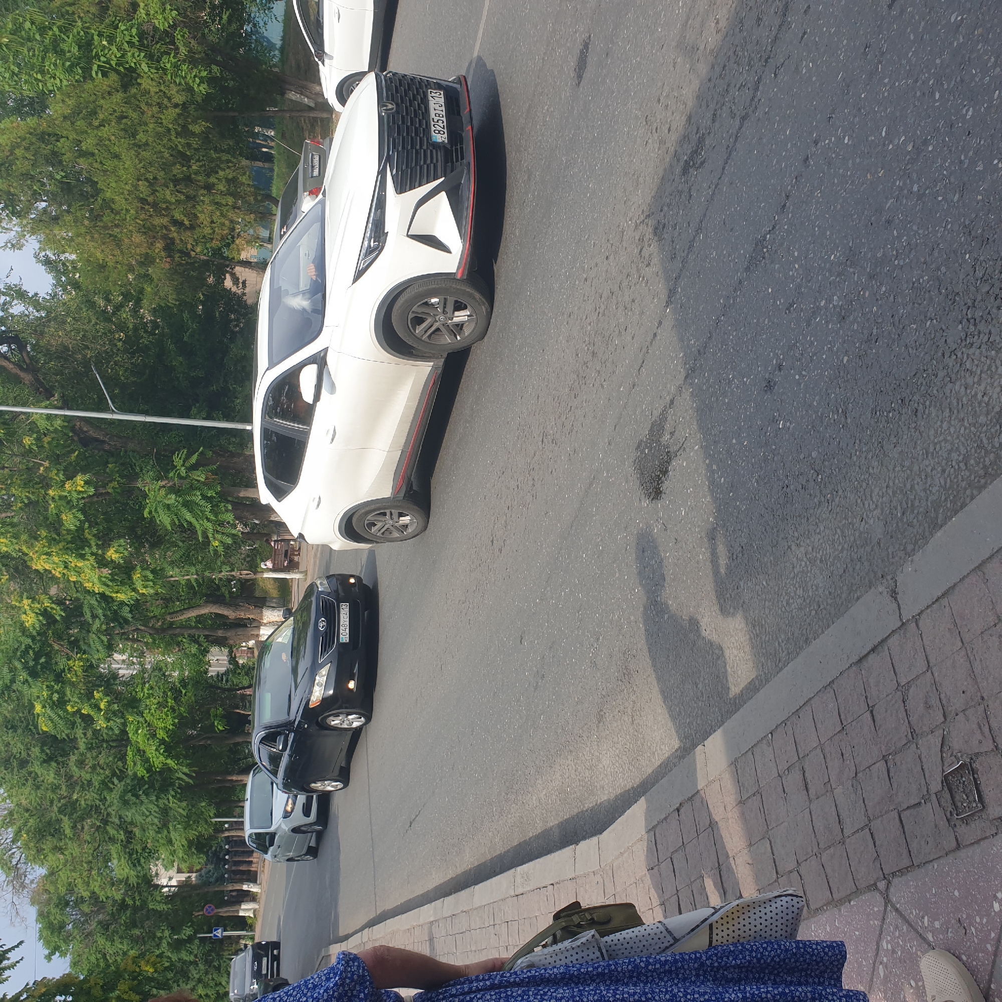 The image shows a street scene in Shymkent, Kazakhstan, with several cars driving on a paved road. The sun is shining, casting shadows on the road. On the left side of the road, a person wearing a blue dress and carrying a bag is standing on a sidewalk made of stone. The road is lined with trees. In the foreground, a white SUV with license plate "KZ 825BIJ 13" is visible. Behind it, a black sedan with license plate "048YCA 13" is also on the road. Other vehicles and trees are visible in the background.