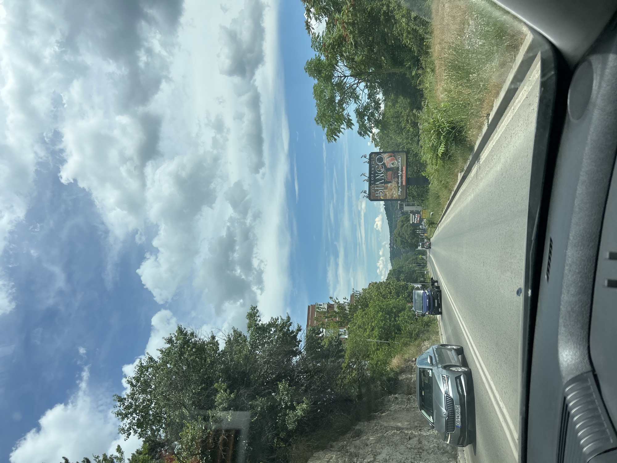 The media file displays a view from inside a car, looking out towards a road and its surroundings. The image is oriented vertically, with the road running diagonally from the bottom right to the upper left.
The scene depicts a paved road with two lanes, marked by white lines. Several cars are visible on the road, traveling in the same direction as the viewer. The car in the foreground, on the right side of the road, is a modern grey sedan with a license plate reading "CB 9438 PC". Further down the road, a blue van is visible, and beyond that, other vehicles are seen in the distance.
The roadside is lined with lush green trees and vegetation. On the left side of the road, there is a steep, rocky embankment covered in some greenery. On the right side, the terrain slopes upwards, also covered with grass and trees.
In the distance, to the left of the road, a multi-story building with a red brick facade is visible behind the trees. Above the trees and to the left of the road, a billboard with an advertisement is prominently displayed. The billboard features text and an image of food, possibly sushi, with the prominent word "BIANCO" in large letters. Below this, there is additional text in a foreign language, which reads "Вірую, нашим стравою!".
The sky is a bright blue, with scattered white, puffy clouds. The sunlight suggests it is daytime, likely midday. The overall impression is of a scenic drive through a verdant area on a clear day. No people are clearly identifiable in the image.