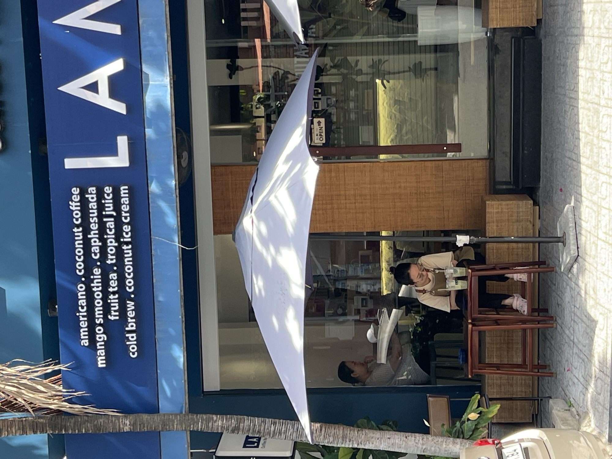 This image captures a street-level view of a cafe in Nha Trang, Vietnam. A blue awning with white lettering is prominently displayed above the storefront, listing menu items such as "americano, coconut coffee," "mango smoothie, caphesuada," "fruit tea, tropical juice," and "cold brew, coconut ice cream." The cafe's name, "LAN," is visible in large white letters on the awning.
In the foreground, a white umbrella provides shade over a seating area. A woman with her hair pulled back is seated at a small wooden table outside the cafe, engaged with her phone. She is wearing a light-colored t-shirt with an apron and dark pants, with pink clogs on her feet. Two glasses with drinks are on the table in front of her. Another woman is visible through the cafe window, seated and looking down. The cafe interior shows displays and shelves. A sign on the umbrella pole reads "OPEN." The street pavement is visible in the foreground with a portion of a light-colored car on the left side of the frame. The overall scene suggests a casual daytime setting at a cafe in Nha Trang.